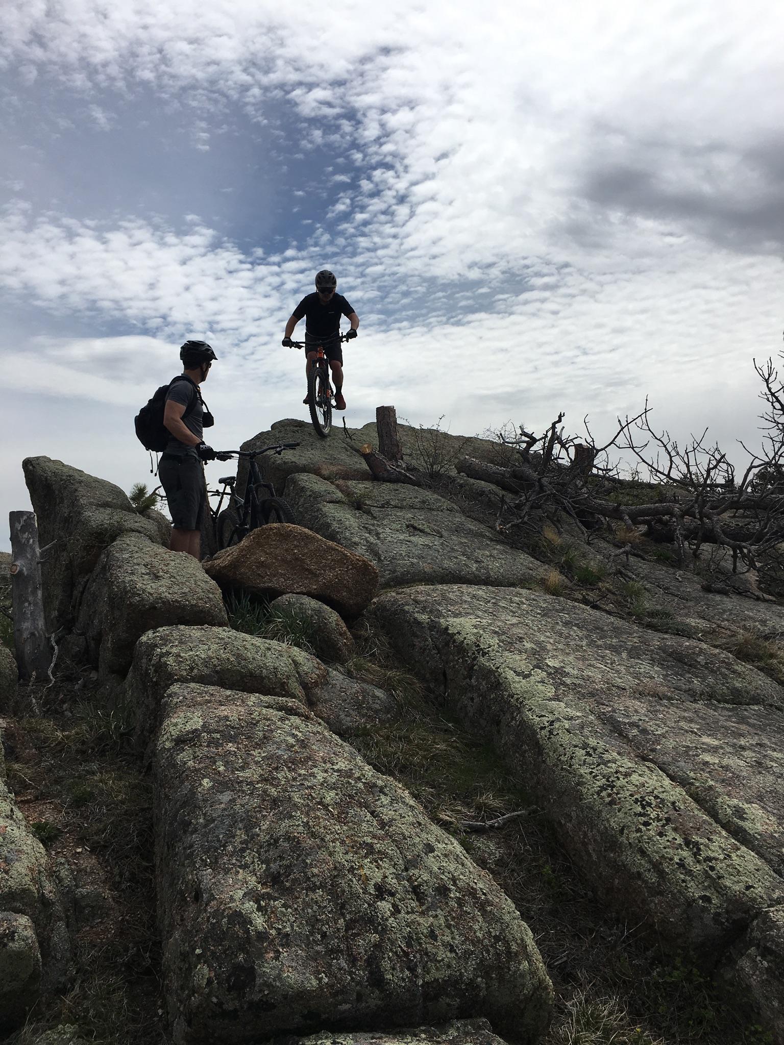 A mountain biker performs a jump over rocky terrain while another biker watches nearby. The scene is set against a backdrop of a cloudy sky, with scattered clouds above and rocky ground covered in moss and grass. Curt Gowdy State Park mountain bike trail.