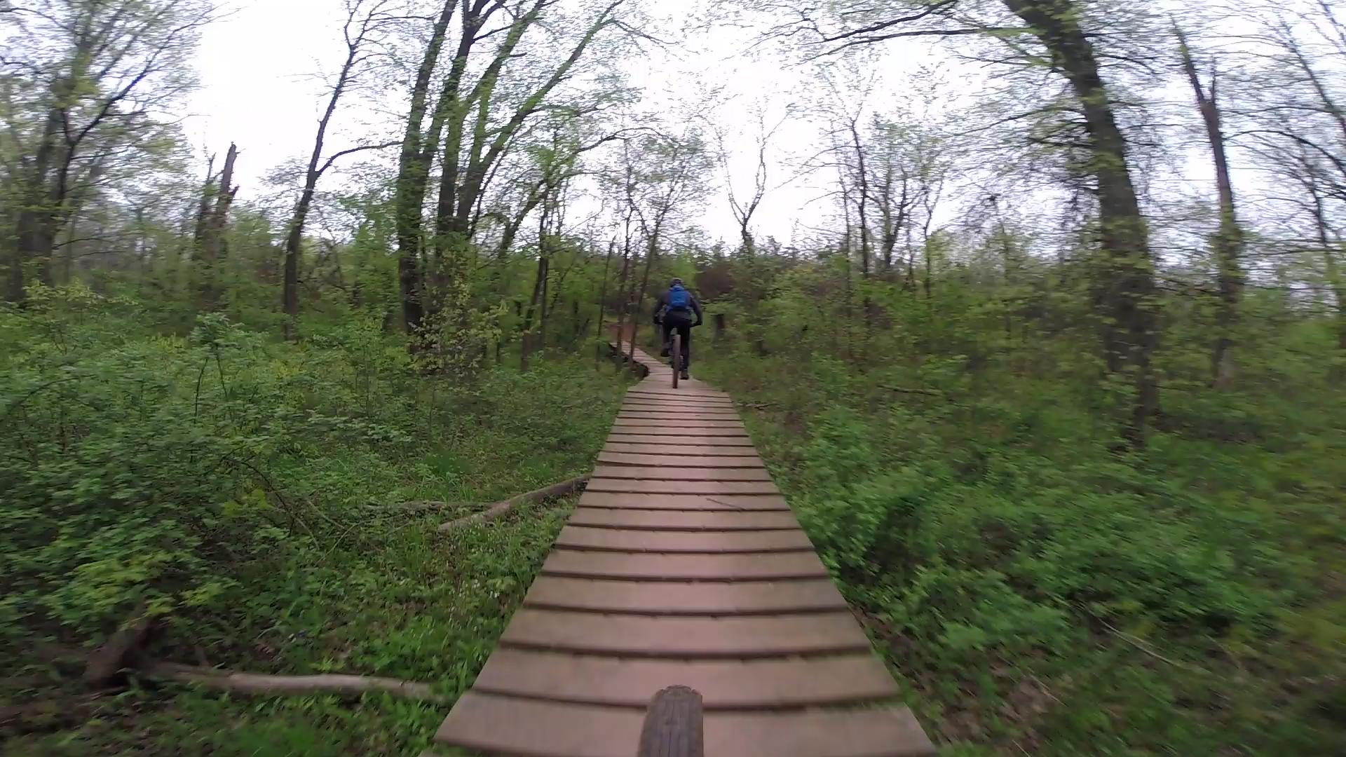 A person riding a mountain bike on a wooden boardwalk trail surrounded by dense green foliage and trees in a forested area on an overcast day. Six Mile Run mountain bike trail.