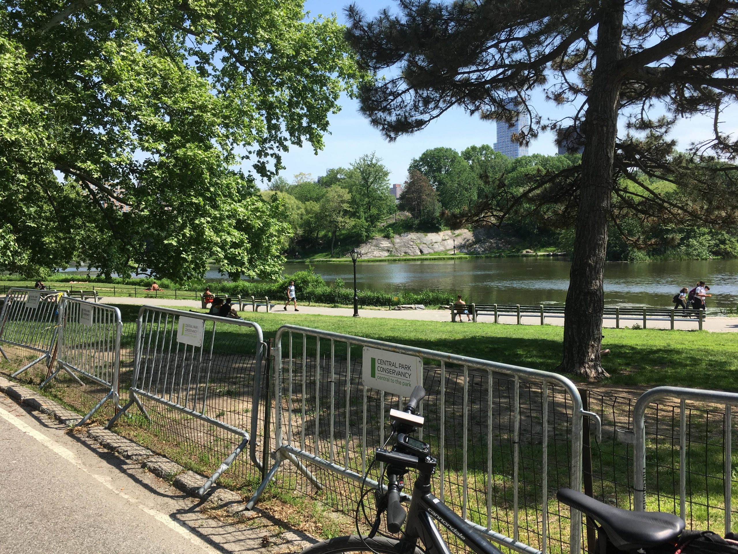 A sunny day in a park featuring a serene lake surrounded by greenery. In the foreground, a bicycle is parked next to security fencing. People can be seen walking and sitting along the pathways by the water, with trees providing shade and a clear blue sky above. Central Park Green way mountain bike trail.