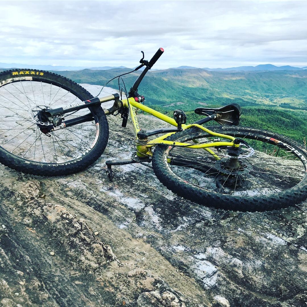 Niner S.I.R. 9: A yellow mountain bike lying on its side on a rocky surface, with a panoramic view of green hills and mountains in the background under a cloudy sky.