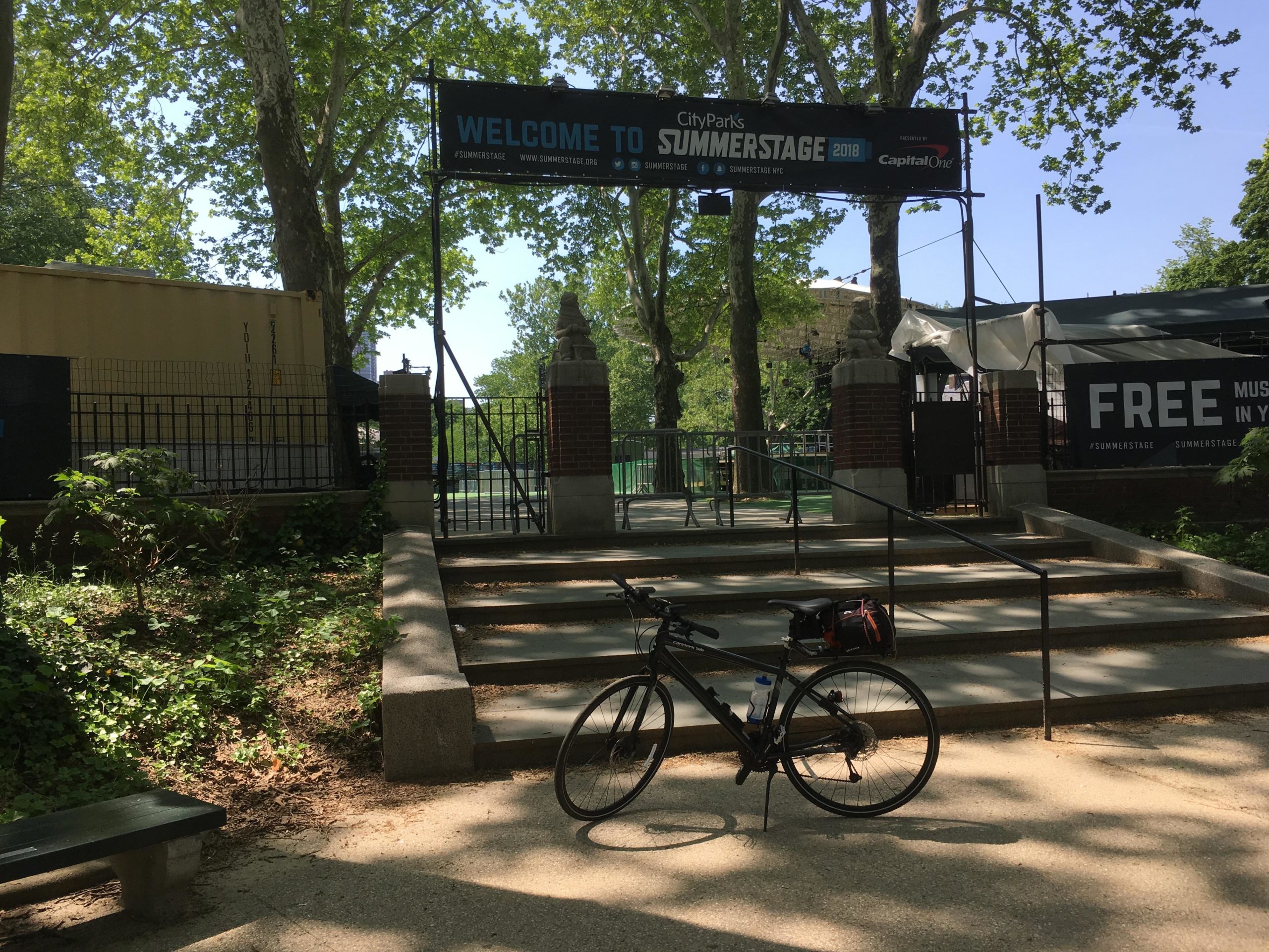 A sunny entrance to City Parks' SummerStage event, featuring a large banner that reads "Welcome to Summerstage 2018." The scene includes steps leading up to the entrance, a parked bicycle on the right, and trees providing shade in the background. There are signs advertising free music events and additional structures visible nearby. Central Park Green way mountain bike trail.
