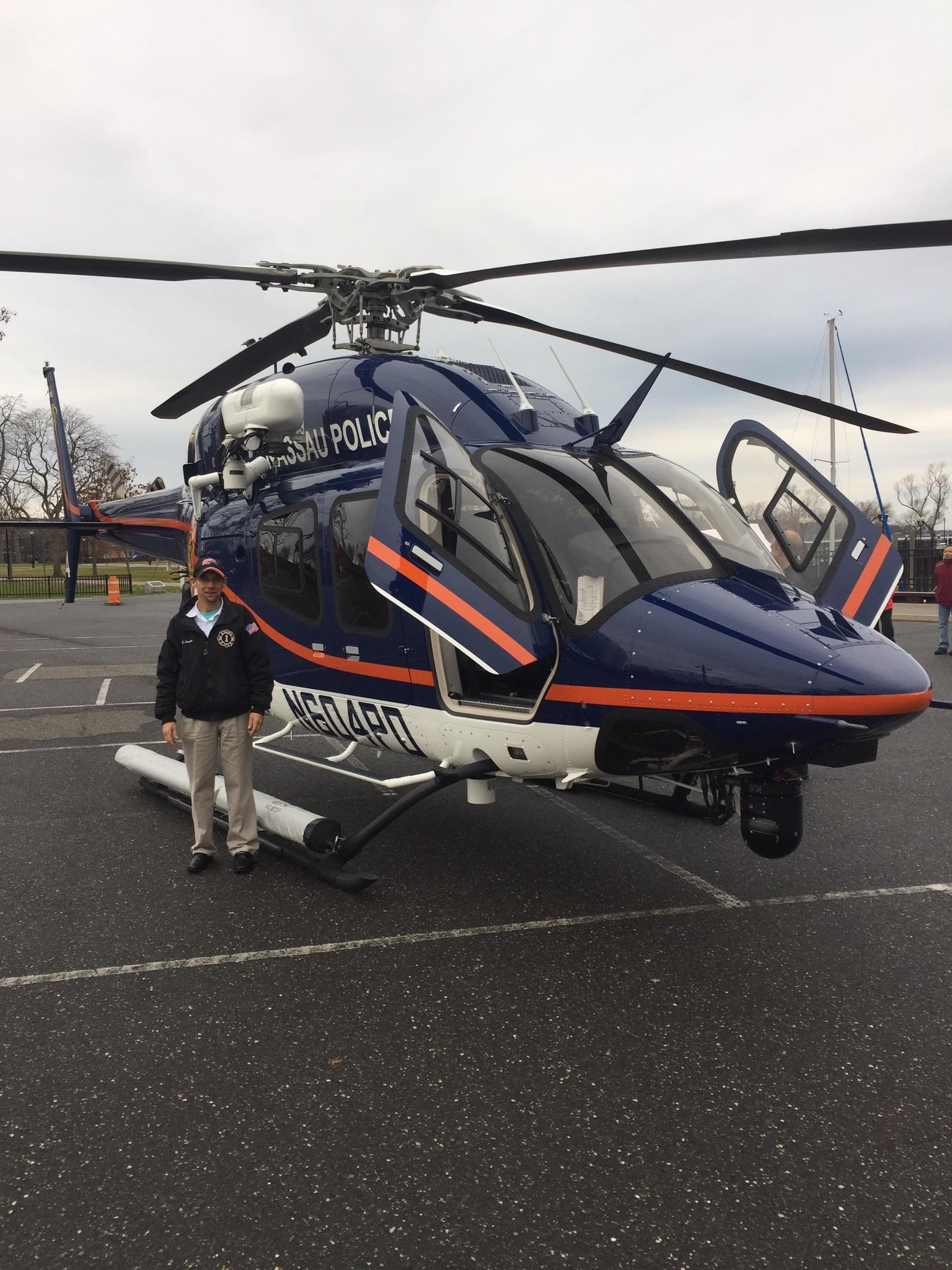 Giant Revel 29er: A police officer stands beside a blue and orange Nassau County Police helicopter, parked on a tarmac. The helicopter features a clear cockpit with partially opened doors and is equipped with surveillance cameras. Overcast skies and a distant view of trees and buildings are visible in the background.