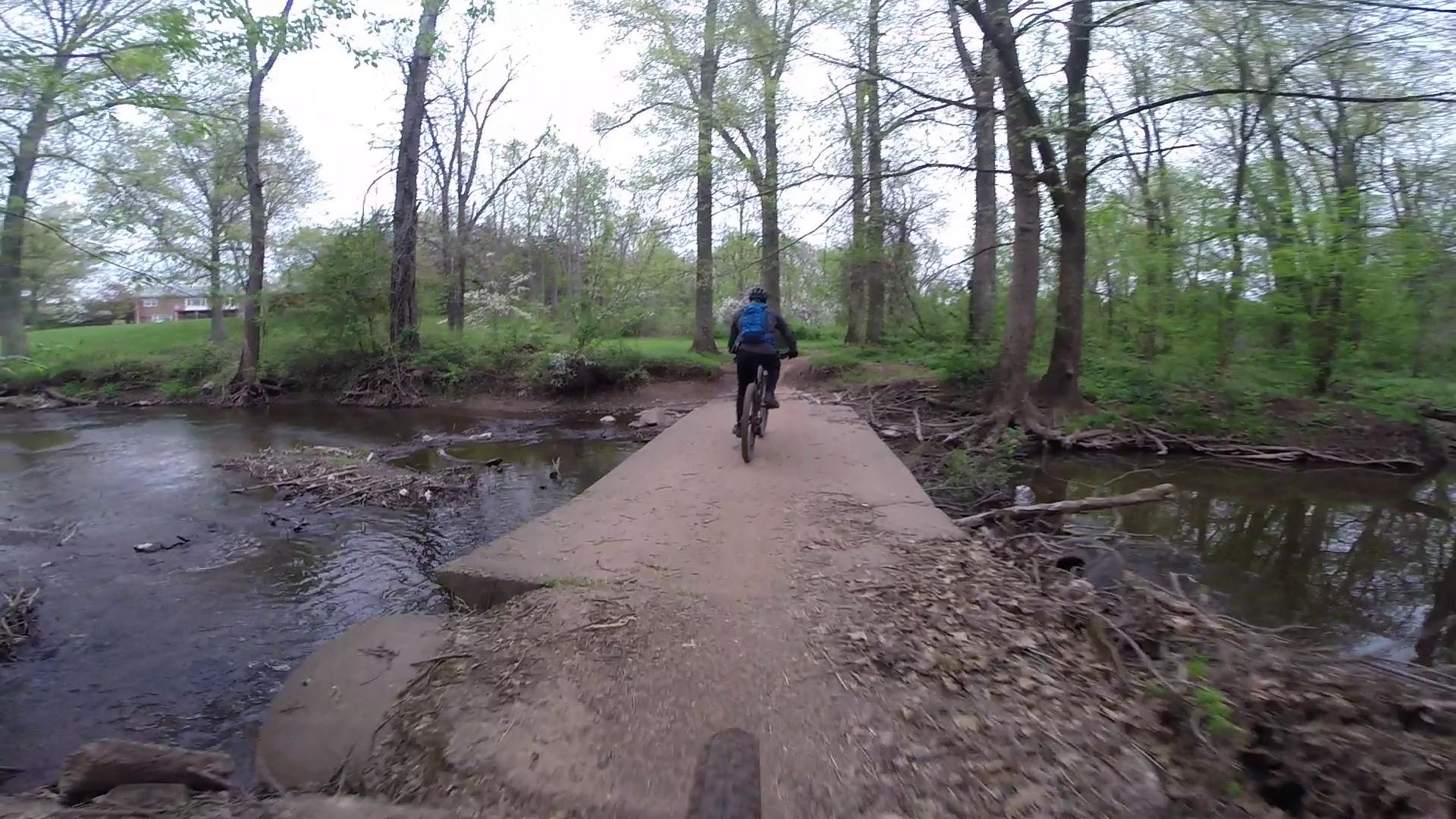 A cyclist riding on a dirt path across a small bridge above a creek, surrounded by lush green trees and foliage. The scene captures a peaceful outdoor setting, ideal for biking and nature exploration. Six Mile Run mountain bike trail.