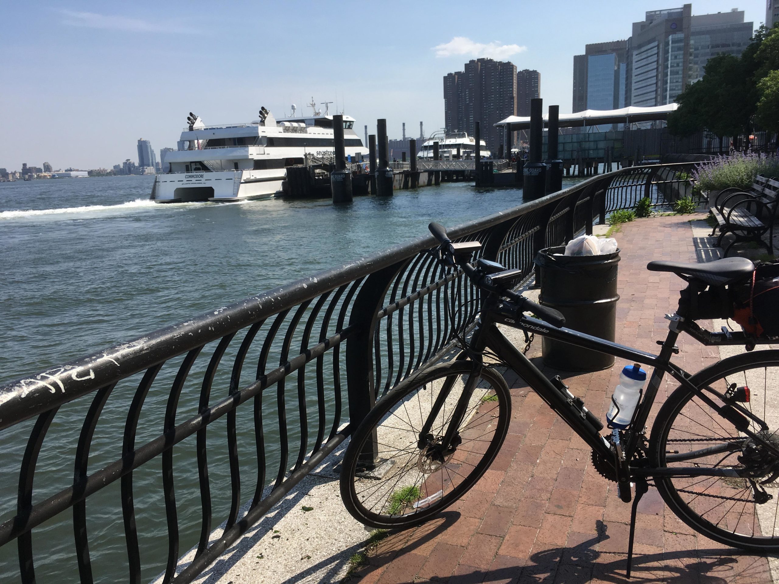 A black bicycle parked along a waterfront promenade with a view of a white ferry approaching the dock. The scene includes a metal railing, a trash bin, and benches in the background, with tall buildings and clear blue skies visible in the distance. East Side Green way 34th st to the Staten Island Ferry mountain bike trail.