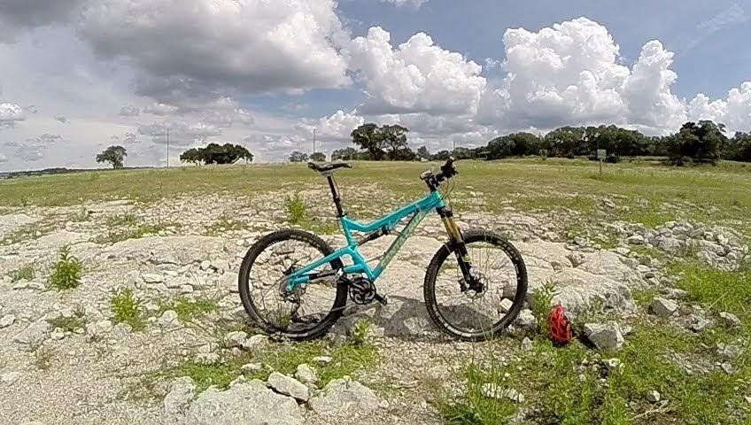 Santa Cruz Heckler: A blue mountain bike parked on rocky terrain, surrounded by sparse greenery under a cloudy sky. The background features a wide, open space with distant trees and a few tall clouds.