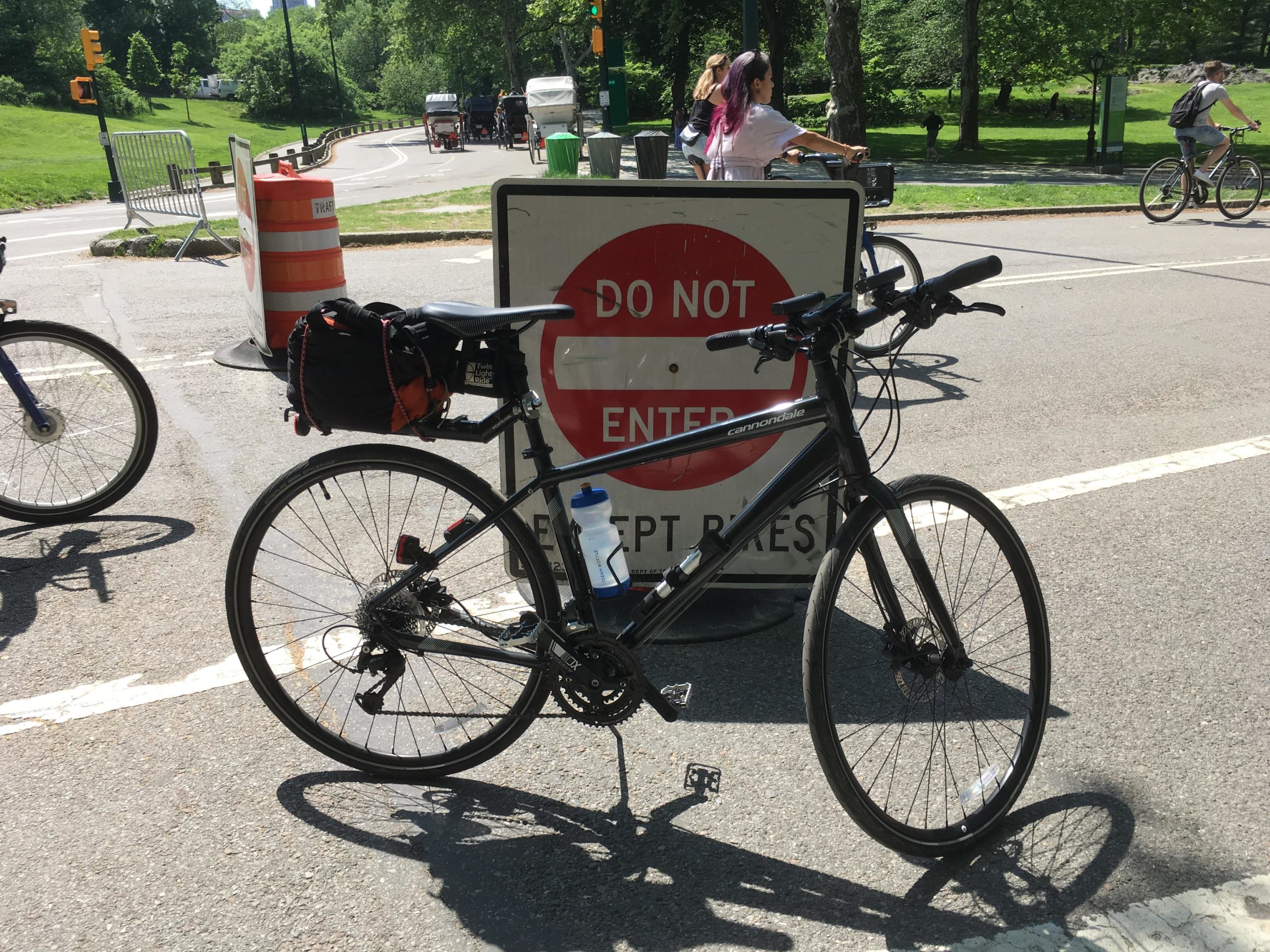 A black bicycle with a backpack and water bottle is parked in front of a "Do Not Enter Except Bikes" sign on a sunny day. In the background, there are other cyclists and a green park area. Road construction barriers are visible nearby, along with trees and a bike path. Central Park Green way mountain bike trail.