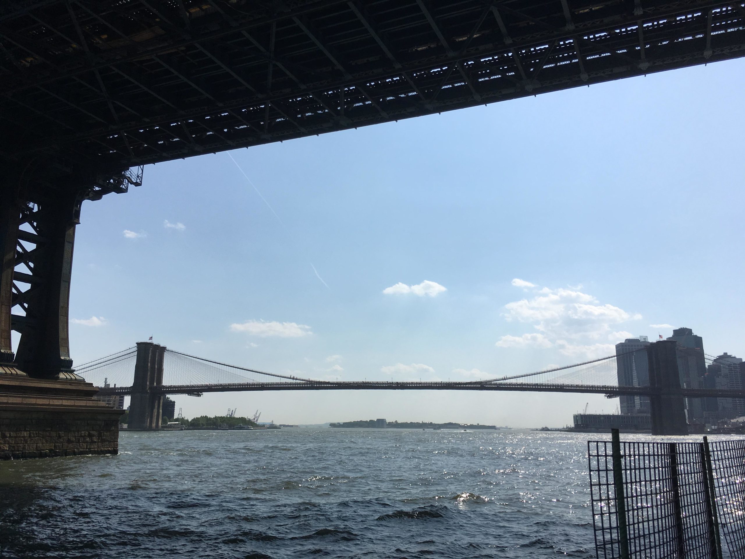 View of the Brooklyn Bridge from beneath another bridge, with a clear blue sky and scattered clouds. The water is visible in the foreground, reflecting sunlight, while city buildings can be seen on the right side of the image. East Side Green way 34th st to the Staten Island Ferry mountain bike trail.