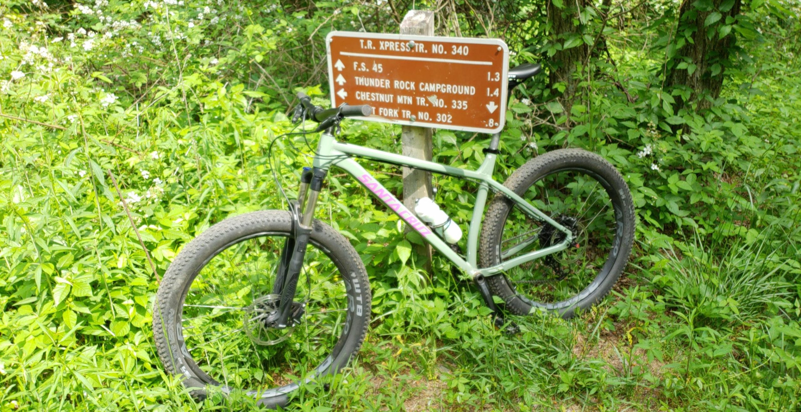 A mountain bike leaning against a trail sign surrounded by lush greenery, indicating directions to various hiking trails and campgrounds. The sign lists distances to F.S. 45, Thunder Rock Campground, Chestnut Mountain Trail, and Fork Trail. Thunderrock Express mountain bike trail.