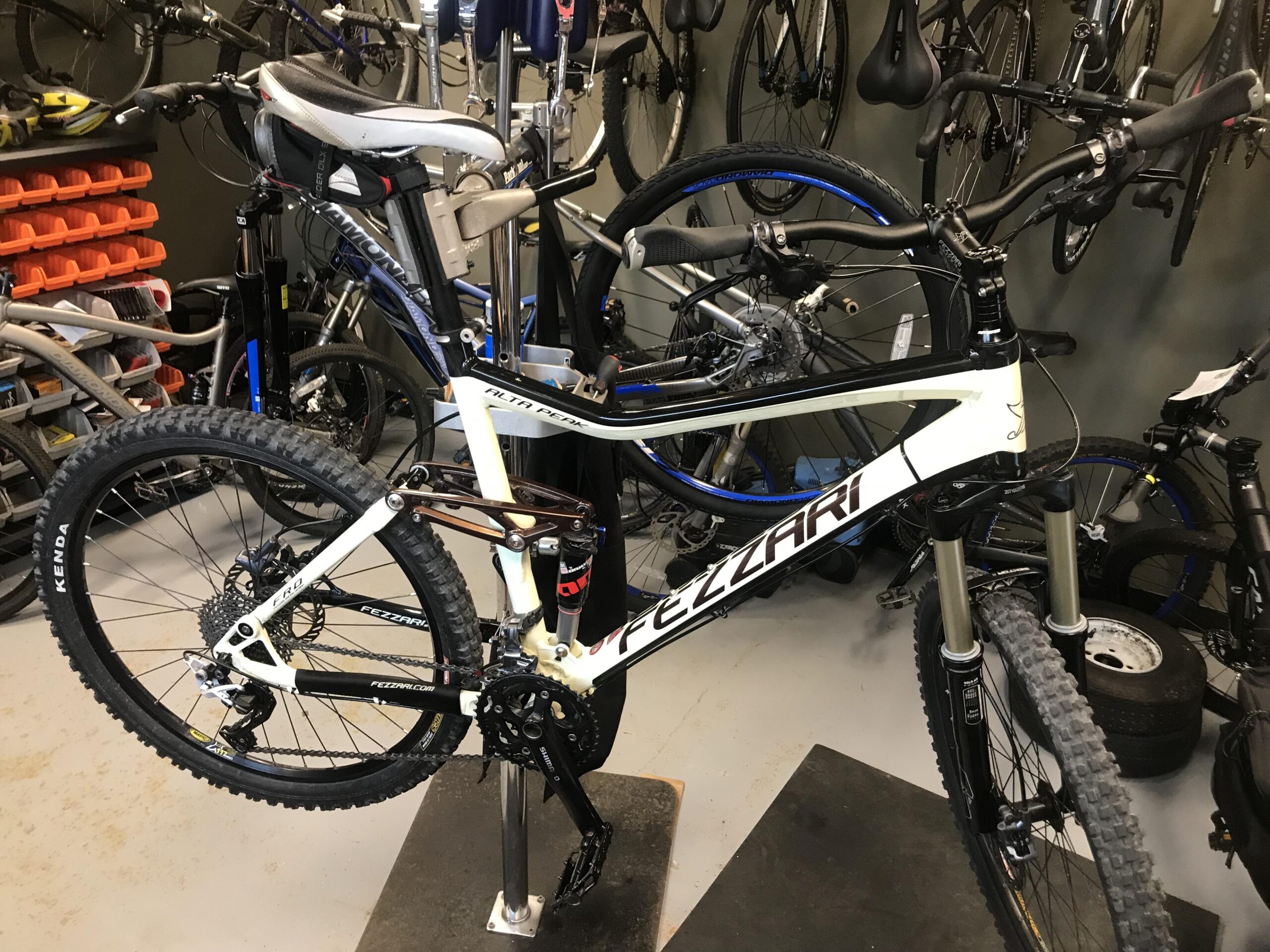 Fezzari Alta Peak: A mountain bike on a maintenance stand in a workshop, surrounded by various other bicycles and cycling equipment. The bike features a white and black frame with the brand name "Fezzari" prominently displayed. The workshop has a clean and organized appearance, with tools and bike parts visible in the background.