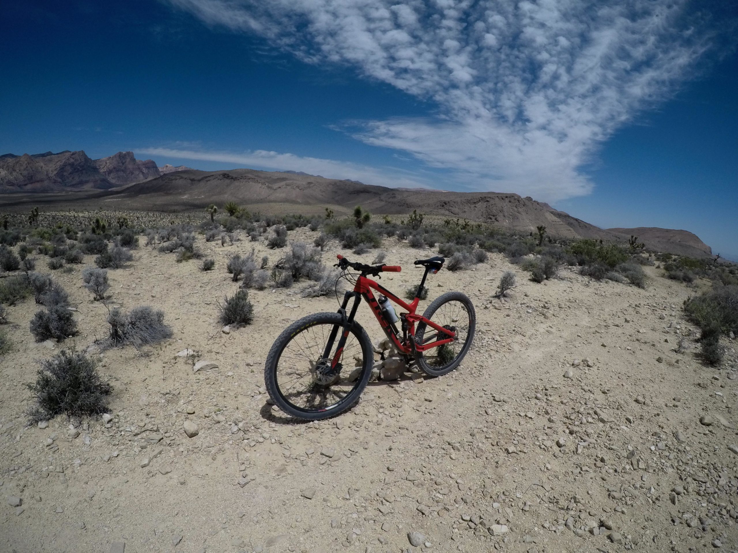 A red mountain bike parked on a dirt trail in a rugged, arid landscape with scrubby vegetation and distant rocky mountains under a clear blue sky with scattered clouds. Blue Diamond mountain bike trail.