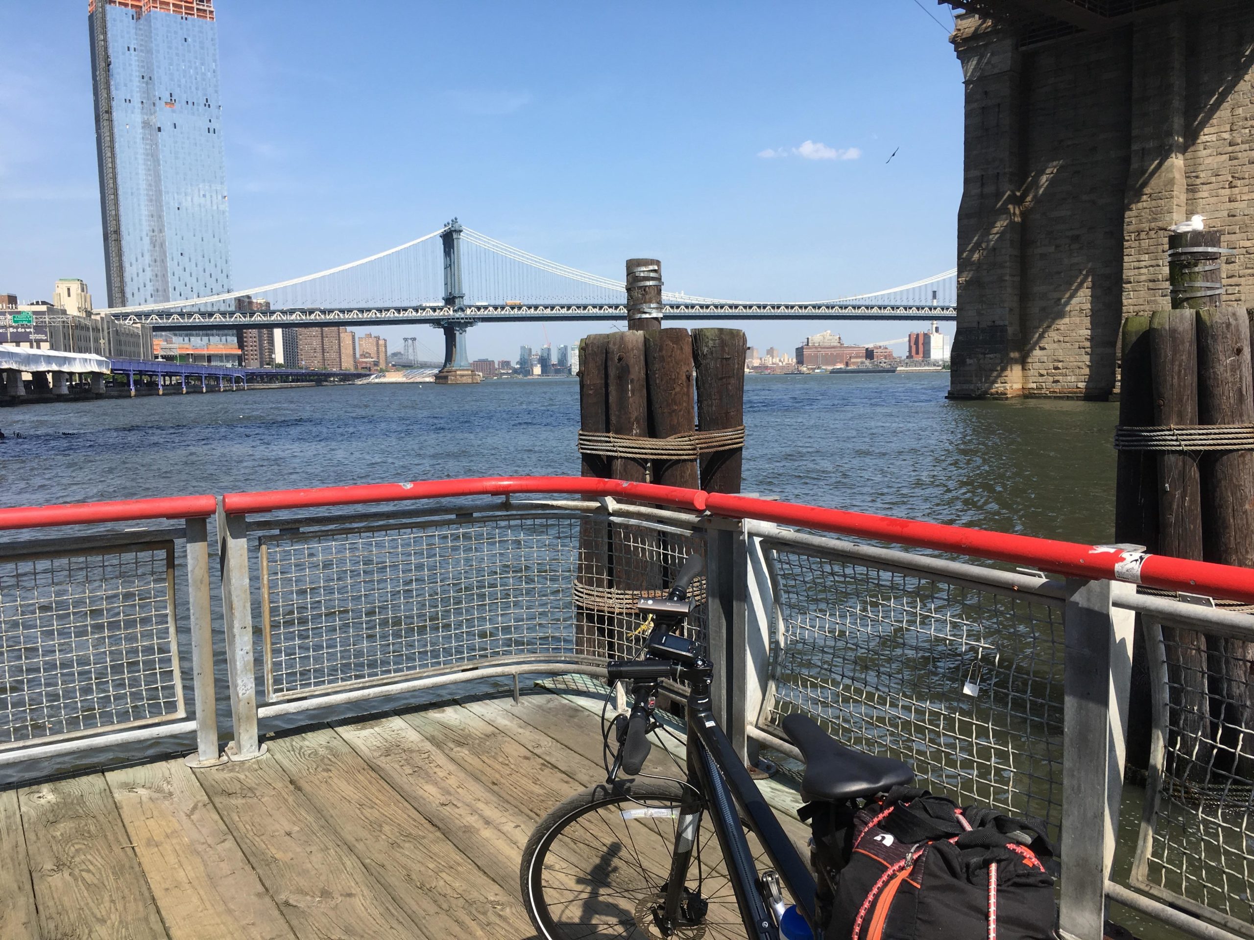 A view of a riverfront scene featuring a bicycle parked near a railing, with the Manhattan Bridge visible in the background. The sky is clear and blue, and the scene captures a mixture of modern buildings and waterfront structures. East Side Green way 34th st to the Staten Island Ferry mountain bike trail.