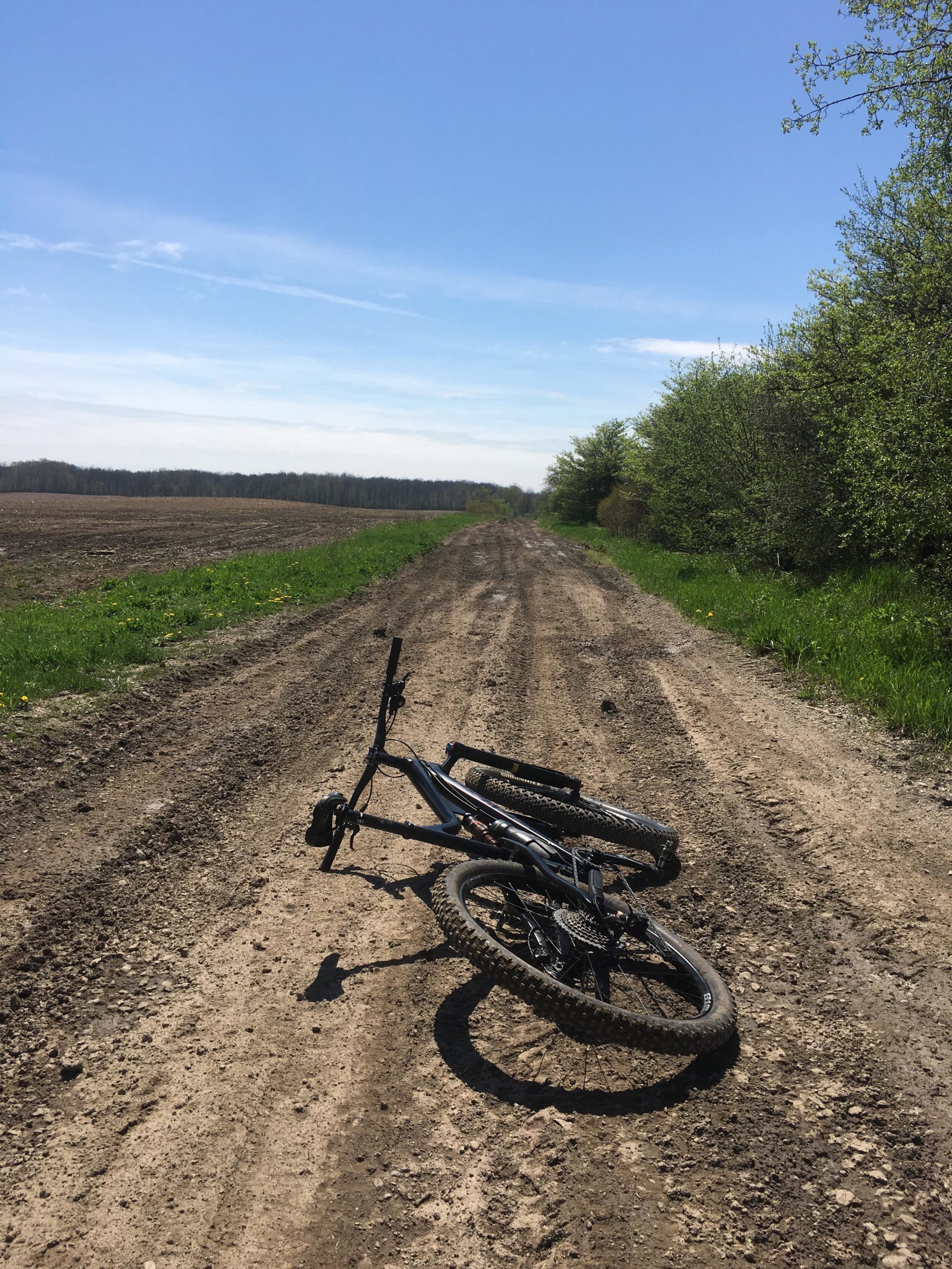 A mountain bike lies on its side on a dirt road flanked by greenery, with fields visible in the background under a clear blue sky. Blind Line mountain bike trail.