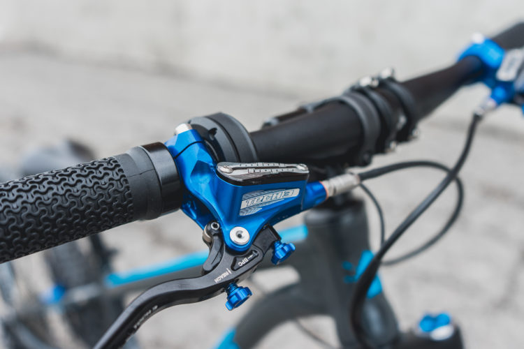 Close-up view of a bicycle handlebar featuring a textured black grip and a blue hydraulic brake lever. The handlebar also shows additional components, including a gear shifter with a marked scale. The background is softly blurred, highlighting the details of the bike's controls.
