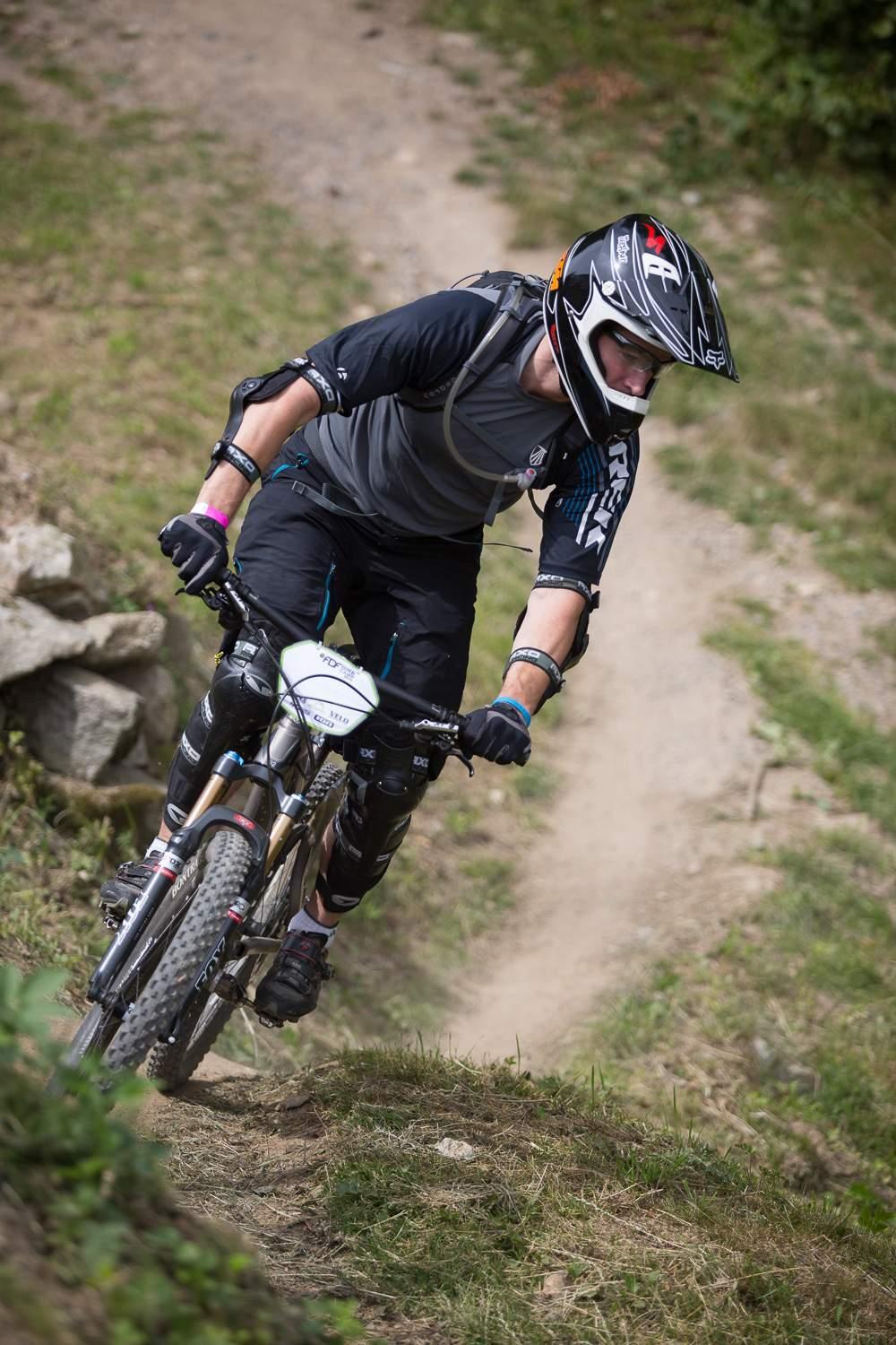 Trek Remedy 9.8 27.5/650b: A mountain biker in protective gear riding down a dirt trail, leaning into the turn. The surrounding landscape features grassy vegetation and scattered rocks, with a blurred background indicating speed and motion.