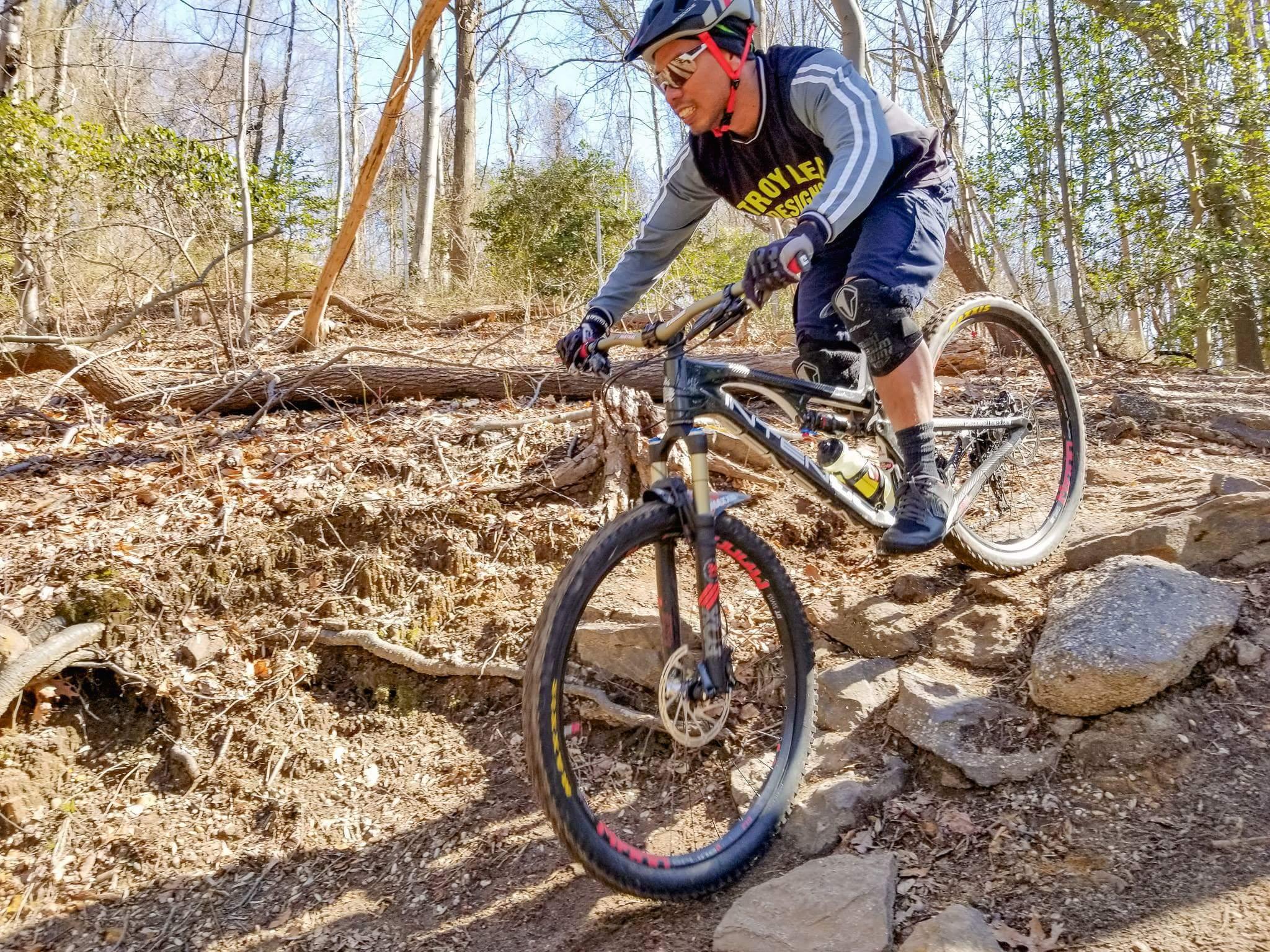 A mountain biker navigating a rocky trail in a forested area, wearing a helmet and protective gear. The rider is focused as they maneuver over stones and uneven terrain, showcasing the challenges of off-road cycling. Hartshorne Woods Park mountain bike trail.