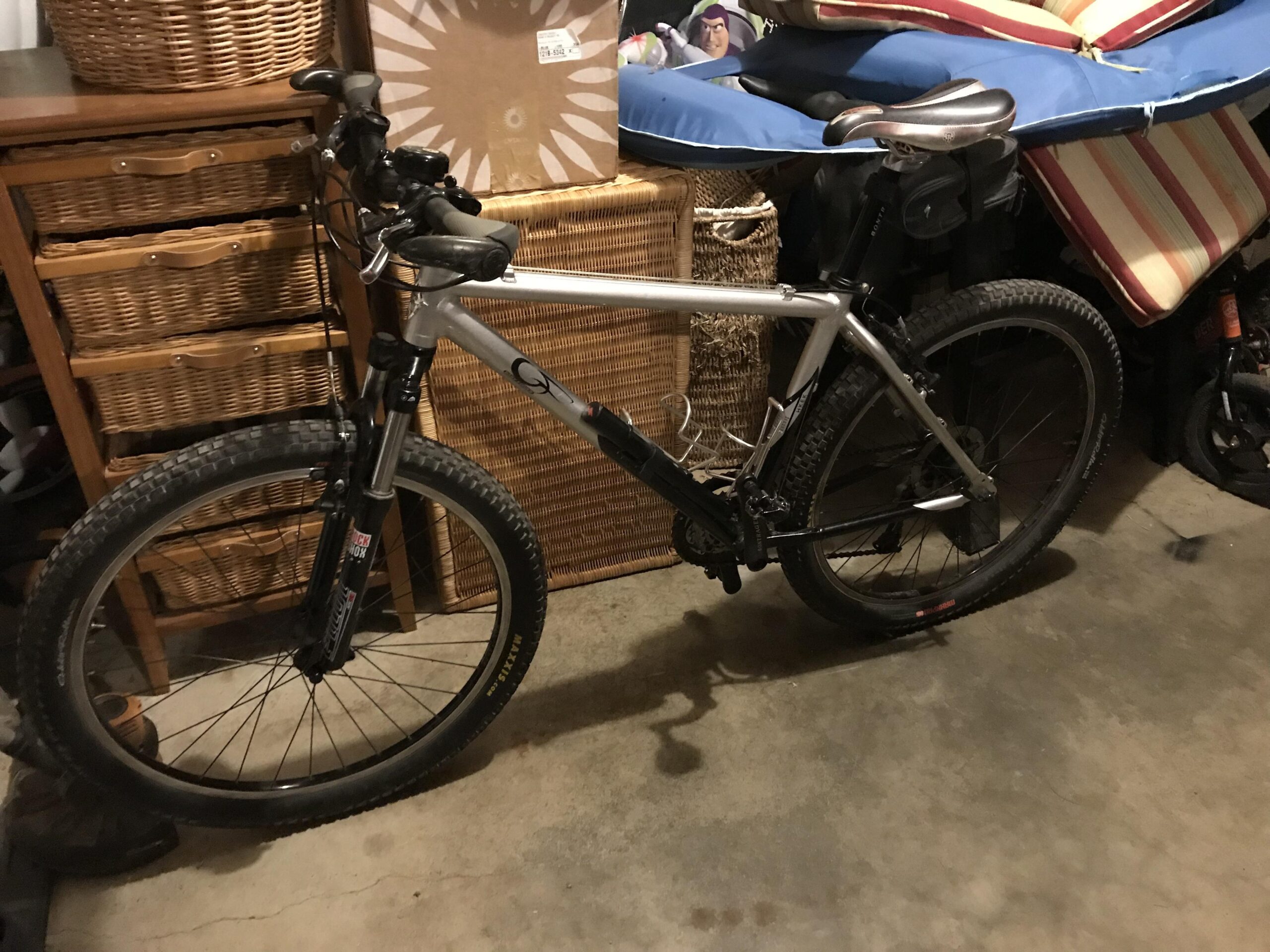 Gary Fisher Hoo Koo E Koo: A silver mountain bike leaning against a wall in a garage, with various storage baskets and items visible in the background.