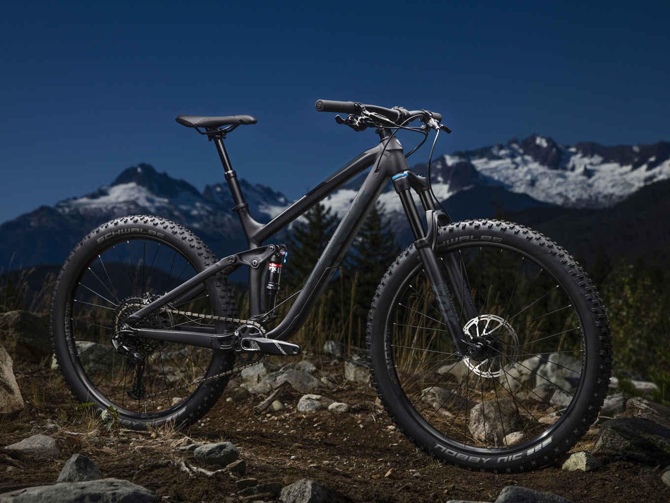Trek Trek Fuel EX 8 27.5 Plus: A black mountain bike is positioned on rocky terrain with a backdrop of snow-capped mountains under a twilight sky. The bike features thick tires, a suspension system, and a sleek frame, emphasizing its rugged design intended for off-road adventures.