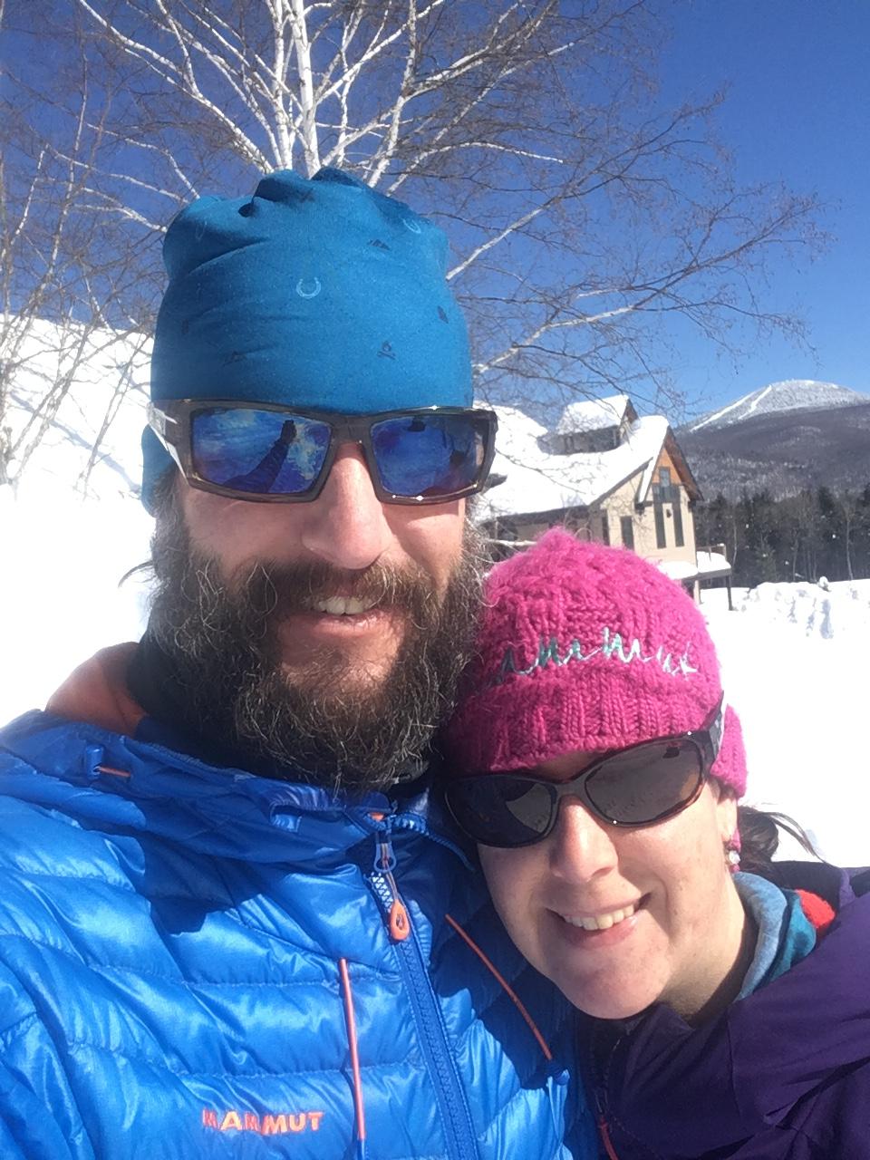 Santa Cruz Hightower 29er/27.5: A happy couple posing for a selfie in a snowy outdoor setting. The man is wearing a blue jacket and headband, has a beard, and sunglasses. The woman is wearing a pink knit hat and a purple jacket, also with sunglasses. Snow-covered mountains and a clear blue sky are visible in the background.