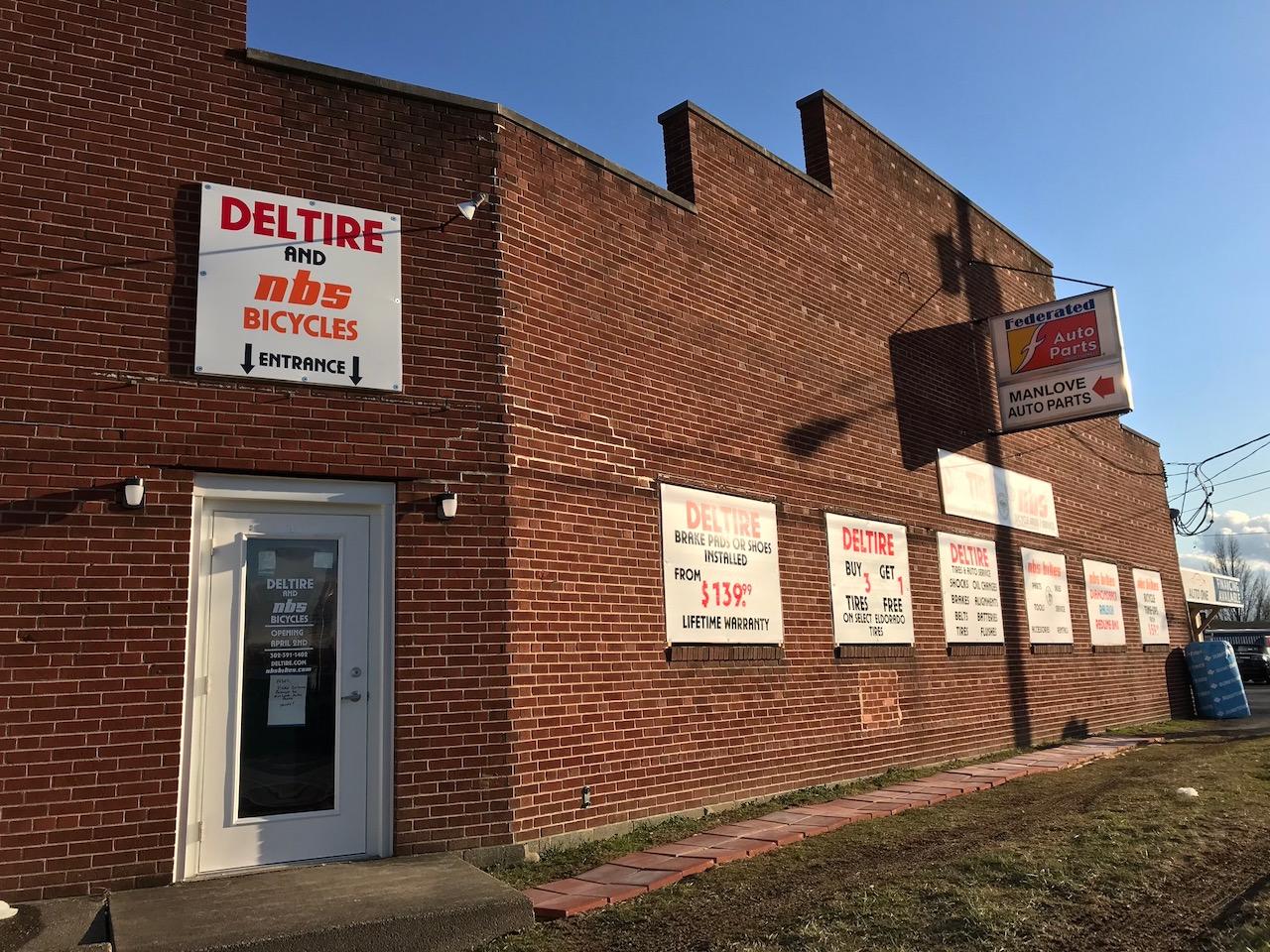 Exterior view of a brick building featuring signs for "Deltire and nbs Bicycles" and "Federated Auto Parts." The entrance door is visible, alongside promotional signs highlighting various bicycle and auto service offers. The image showcases a sunny day, with a clear blue sky in the background.