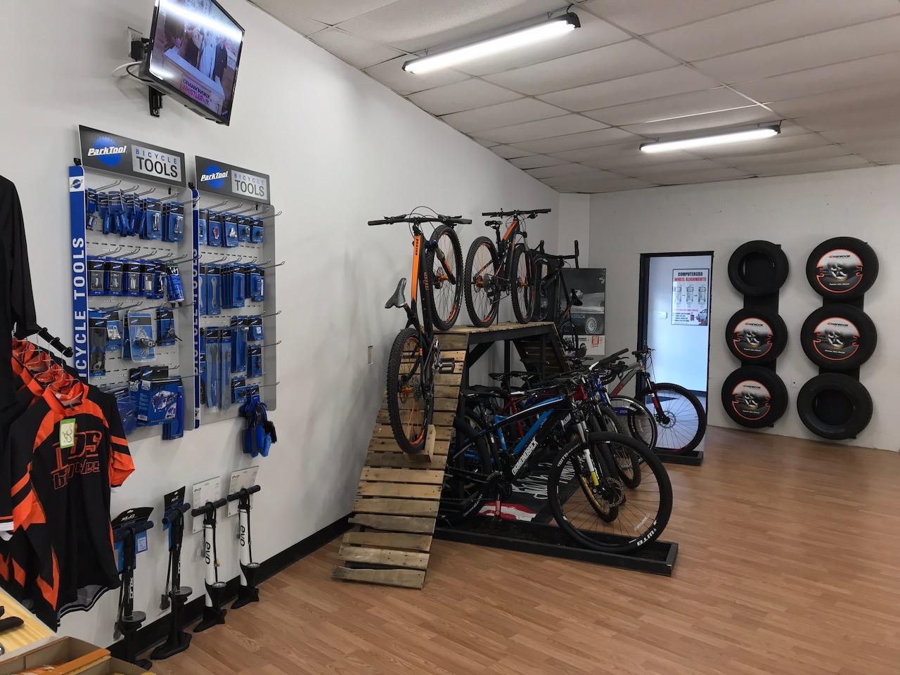 A shop interior featuring a display of bicycle tools on the wall, a wooden ramp with bicycles arranged on it, and additional bicycles on the floor. A television is mounted on the wall, and there are stacked tires displayed nearby. The space has wooden flooring and is well-lit.