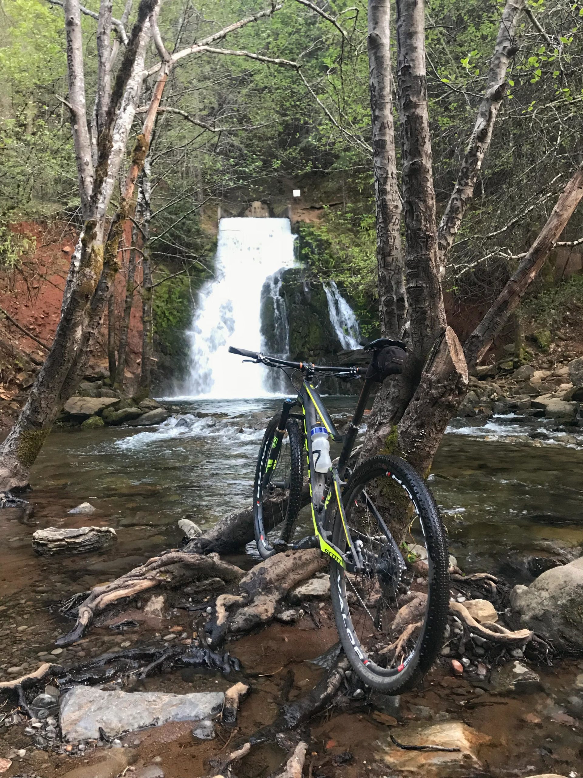 A mountain bike stands near a flowing waterfall, surrounded by lush greenery and water. The bike is positioned on rocky terrain and partially submerged roots, creating a serene natural setting. The cascading water in the background adds to the tranquil atmosphere of the scene. Fleming Meadows mountain bike trail.