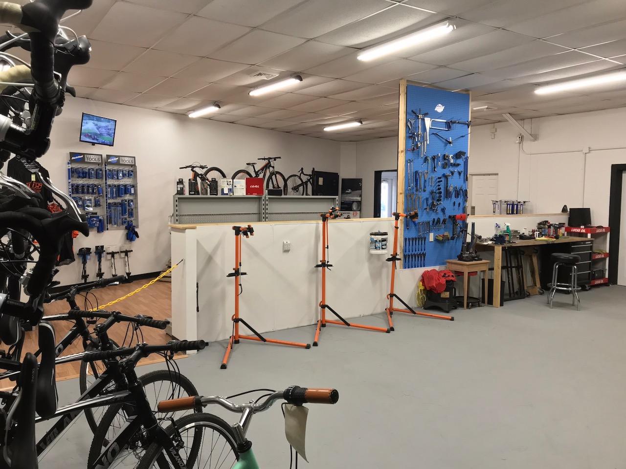 A well-organized bicycle repair shop interior featuring multiple bikes on display, a wall-mounted TV, and various tools and accessories for bike maintenance. The workspace includes orange repair stands and a wooden table with additional equipment, all set against a clean, bright environment with a gray floor and overhead lighting.