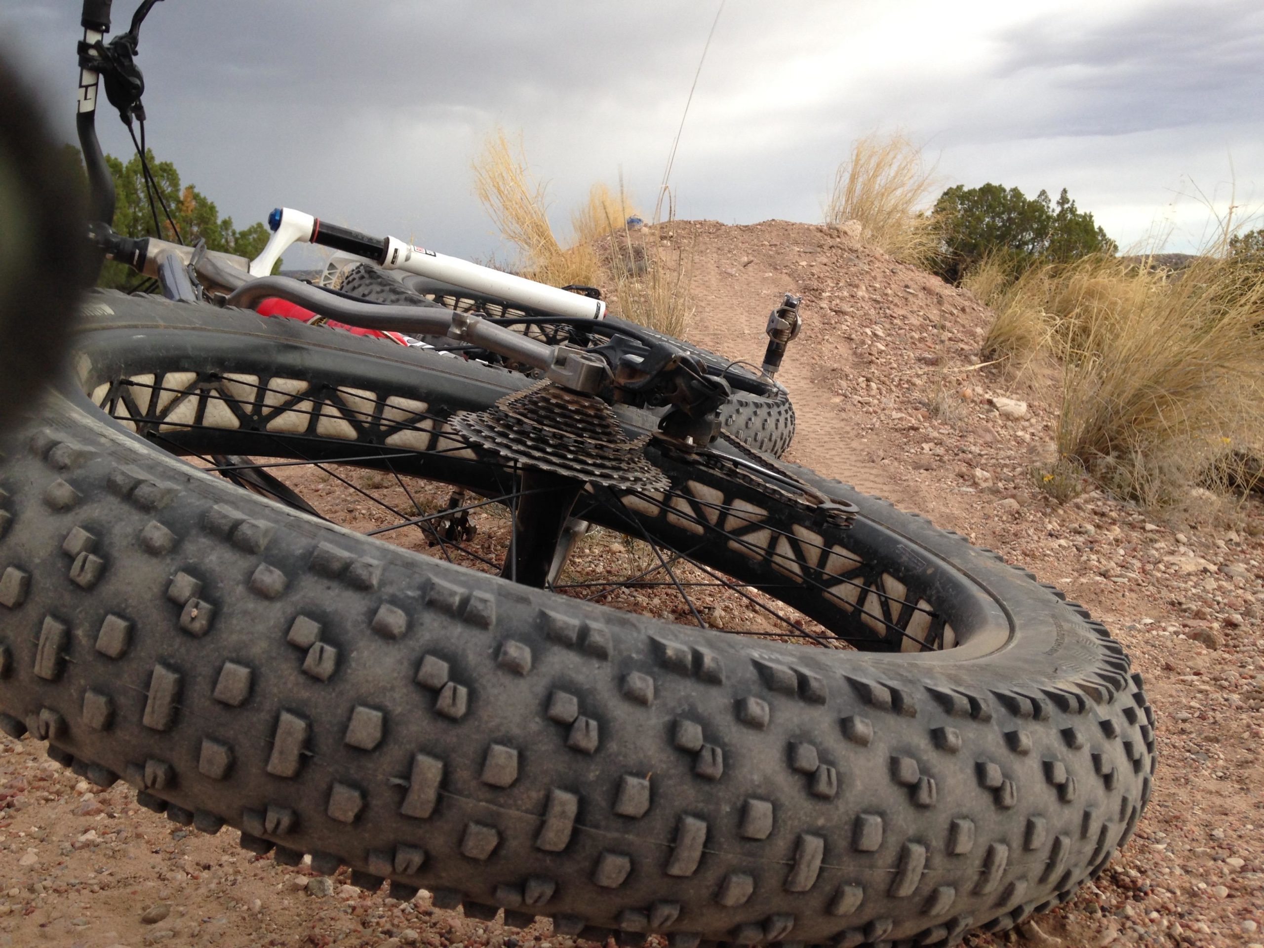 Close-up view of two mountain bike tires resting on a gravel path with sparse vegetation and a cloudy sky in the background. Dangerous D's Jump Line mountain bike trail.