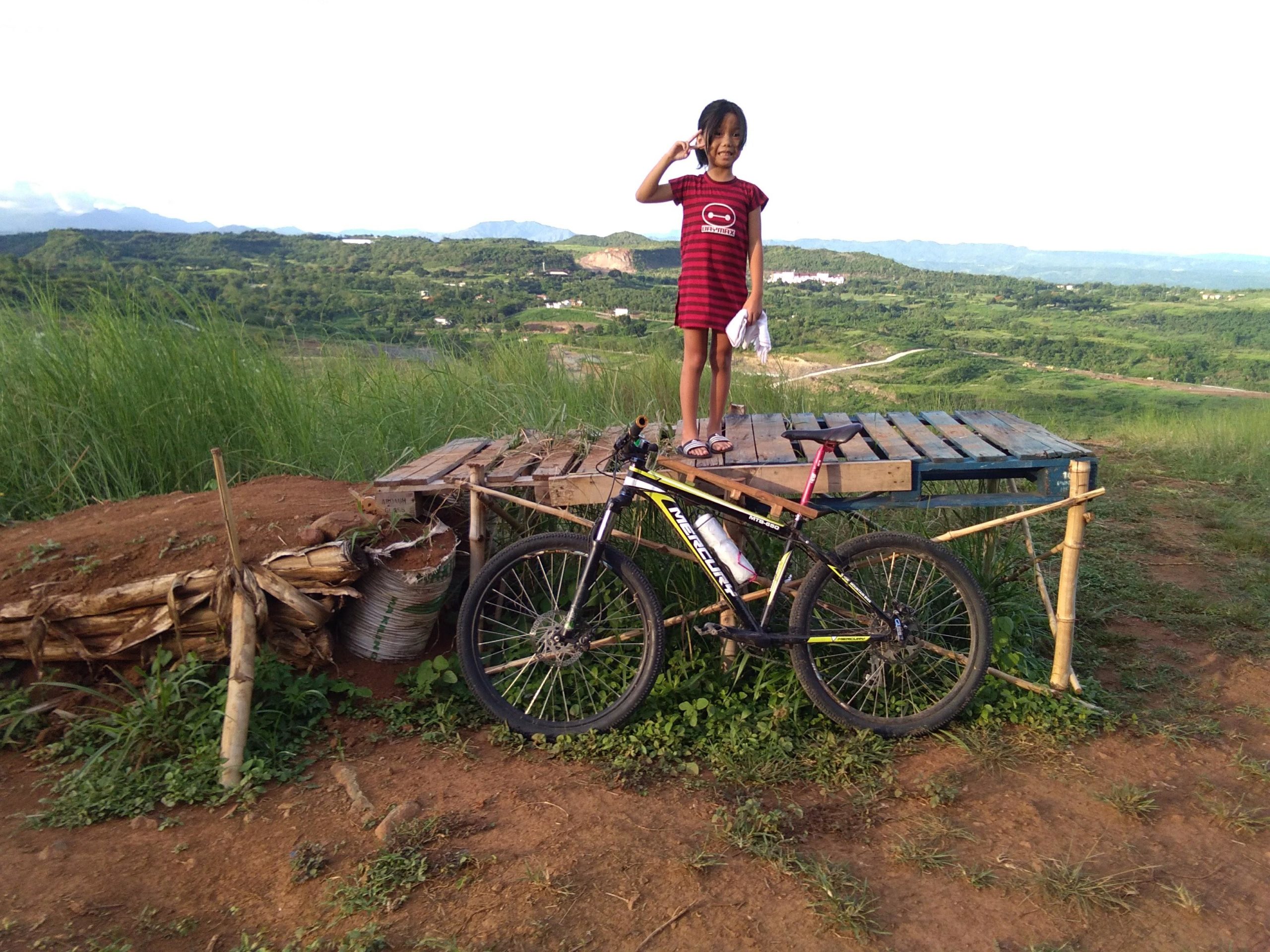 A young girl in a red striped dress stands on a wooden platform, smiling and waving. She is beside a mountain bike on a dirt path, with lush green hills and a clear sky in the background. Pag-asa Antena mountain bike trail.