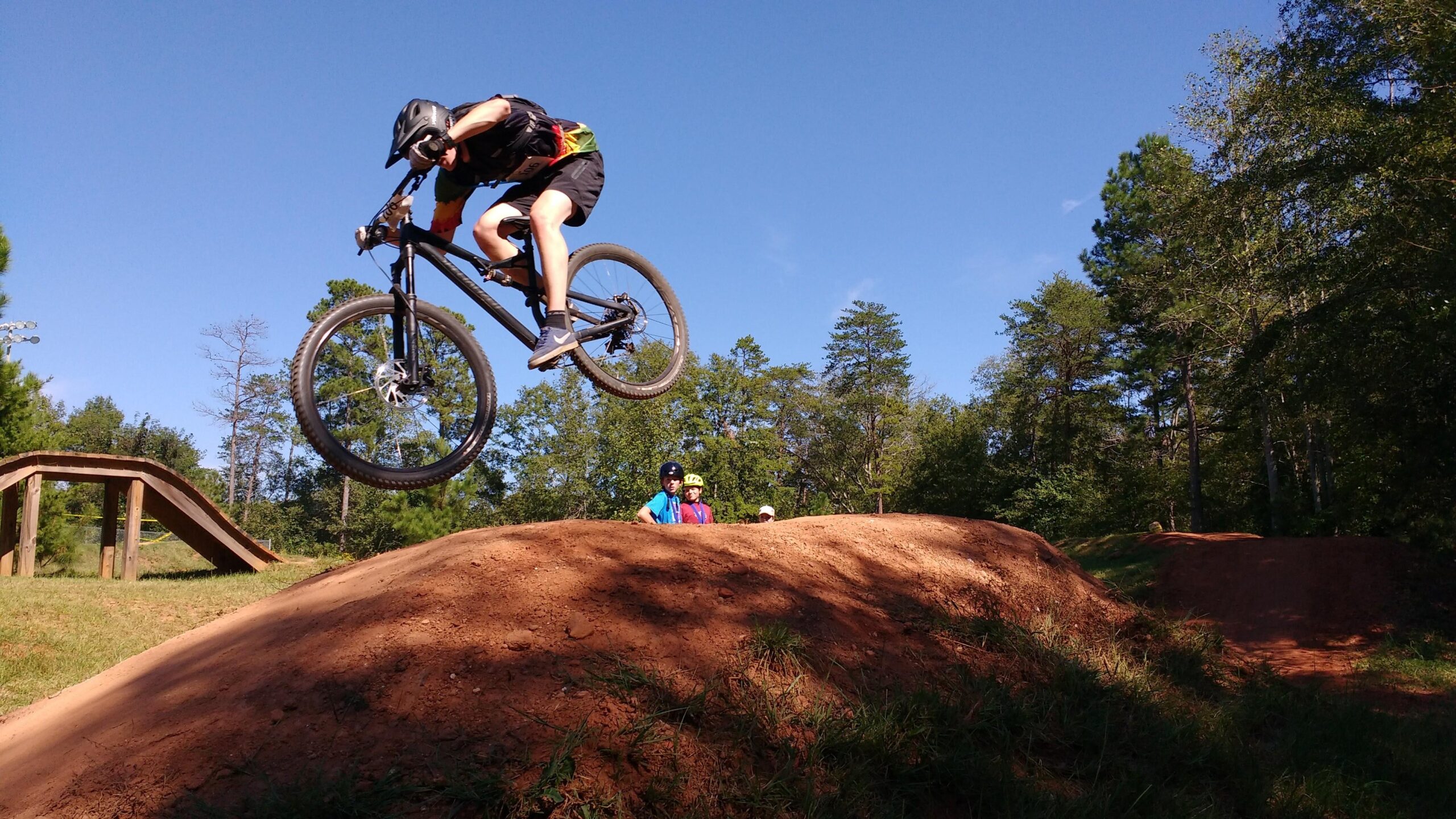 Specialized Stumpjumper FSR 29: A mountain biker in a helmet and jersey performs a jump above a dirt ramp, with two spectators watching from the side. Lush trees and a clear blue sky provide a backdrop, indicating a sunny day at a biking park.