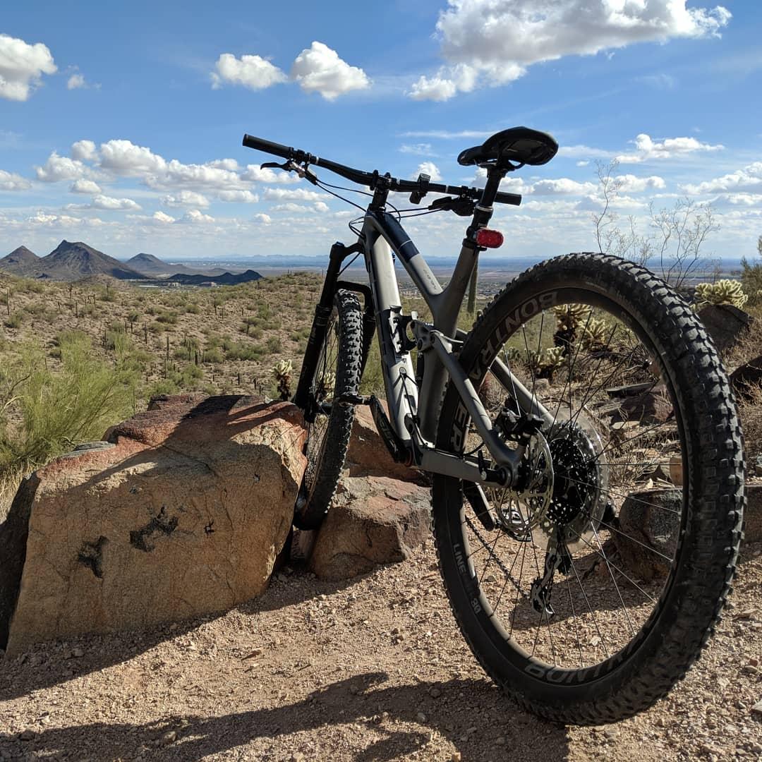Trek Fuel EX 9.8: A mountain bike resting on a rock in a desert landscape, with rugged mountains in the background and a blue sky featuring fluffy white clouds. The scenery is dotted with cacti and shrubs typical of arid environments.