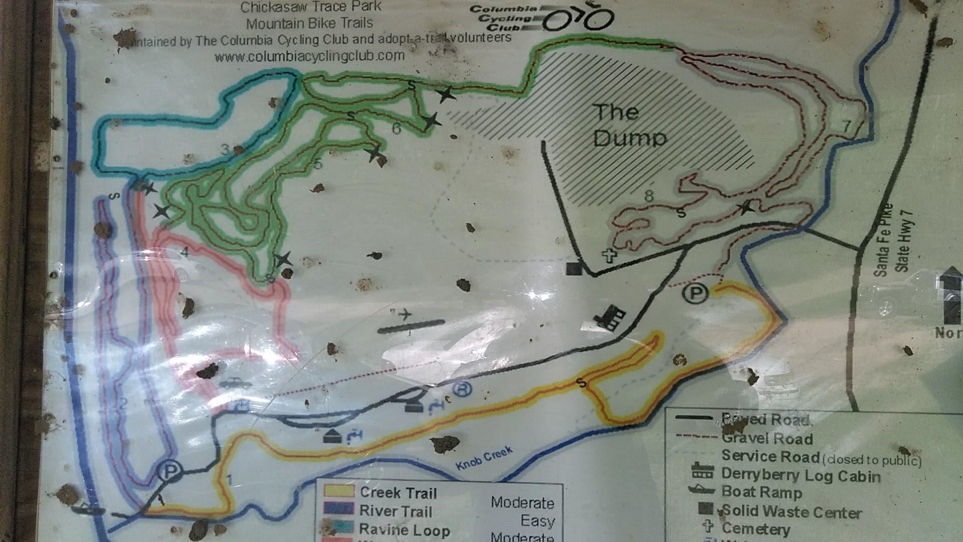 Map of Chickasaw Trace Park mountain bike trails, showing various trails marked in different colors: blue for the creek trail, yellow for the river trail, and pink for the ravine loop. Key features such as parking areas, a boat ramp, and local landmarks are also indicated. The map is slightly worn and has minor dirt marks. Chickasaw Trace mountain bike trail.