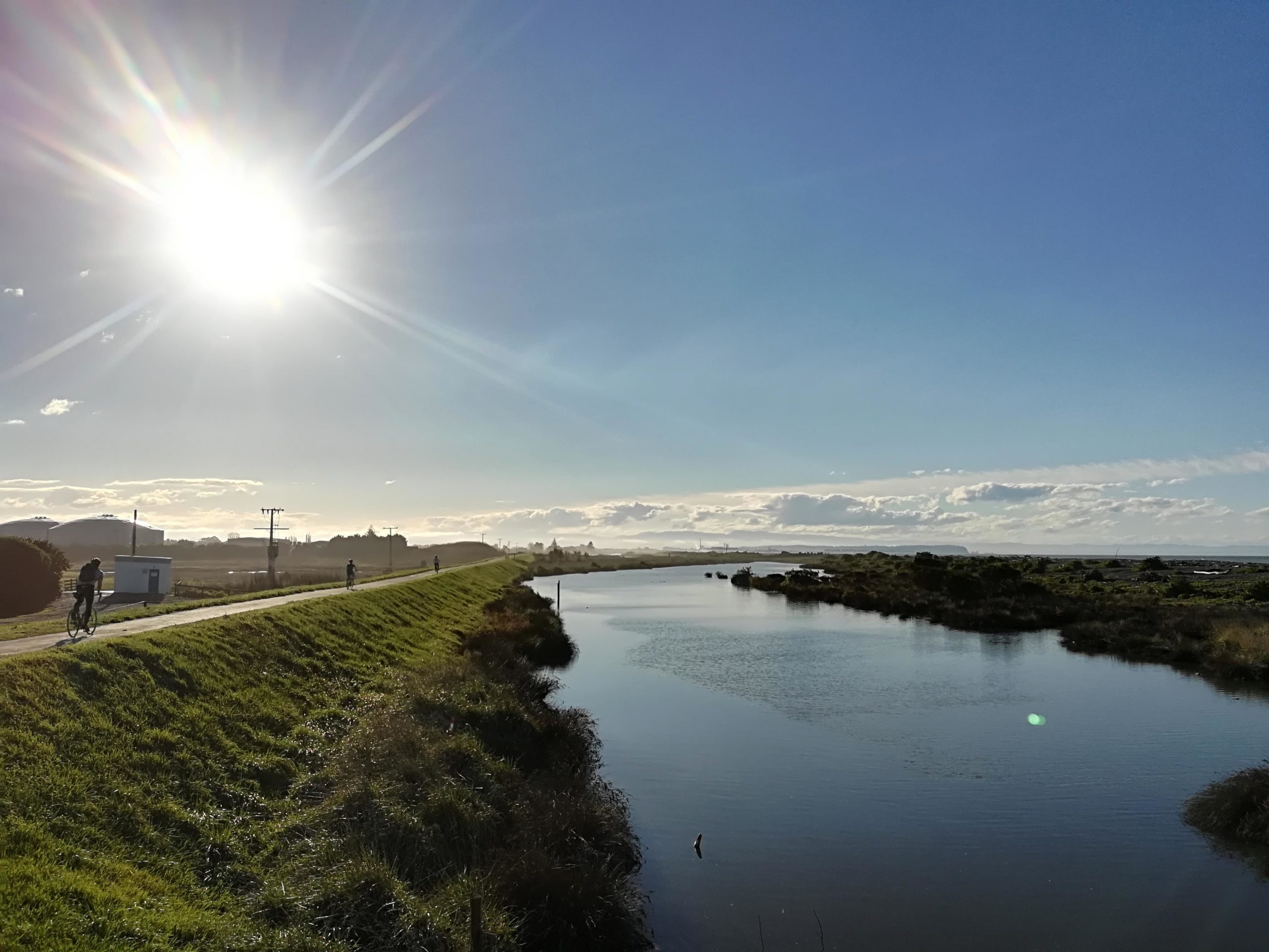 A scenic view of a canal bordered by lush green grass, with a clear blue sky and bright sun shining overhead. In the background, soft hills and distant buildings are visible. A pathway runs alongside the water, where a few cyclists can be seen riding. The tranquil water reflects the sky, creating a peaceful outdoor environment. Hawke