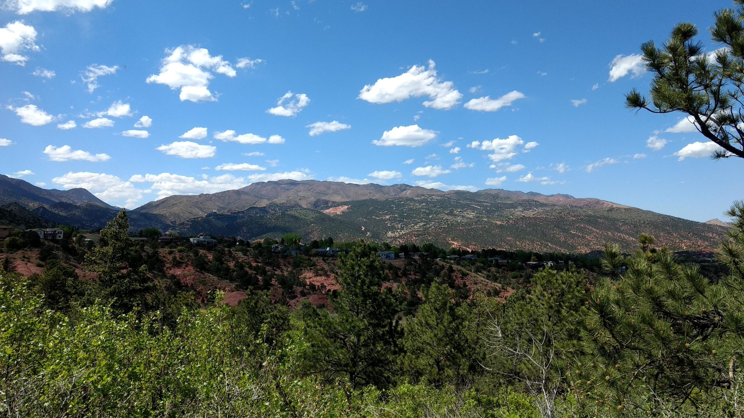 A scenic view of rolling mountains under a blue sky dotted with white clouds. The foreground features lush green trees and shrubs, while the background showcases mountainous terrain with varying shades of brown and green. Small residential buildings are visible nestled among the trees on the hillside. Red Rock Canyon mountain bike trail.