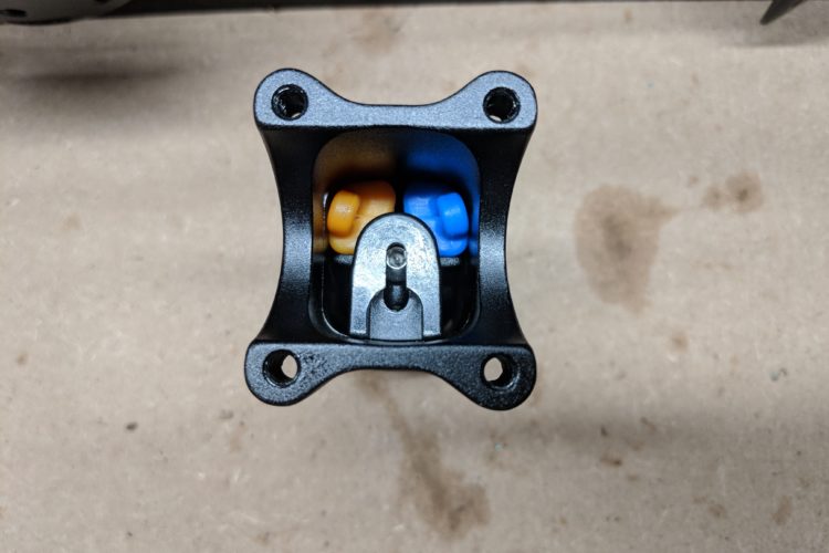 A close-up view of a black mechanical component with a central lever and colored plastic elements (yellow and blue) visible inside. The component is mounted on a flat, light-colored surface, likely a workbench.