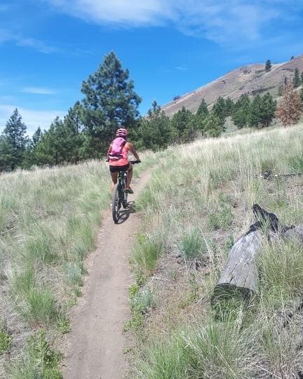 A person riding a mountain bike on a dirt trail surrounded by grass and trees, with a blue sky and hills in the background. Kenna Cartwright Park mountain bike trail.