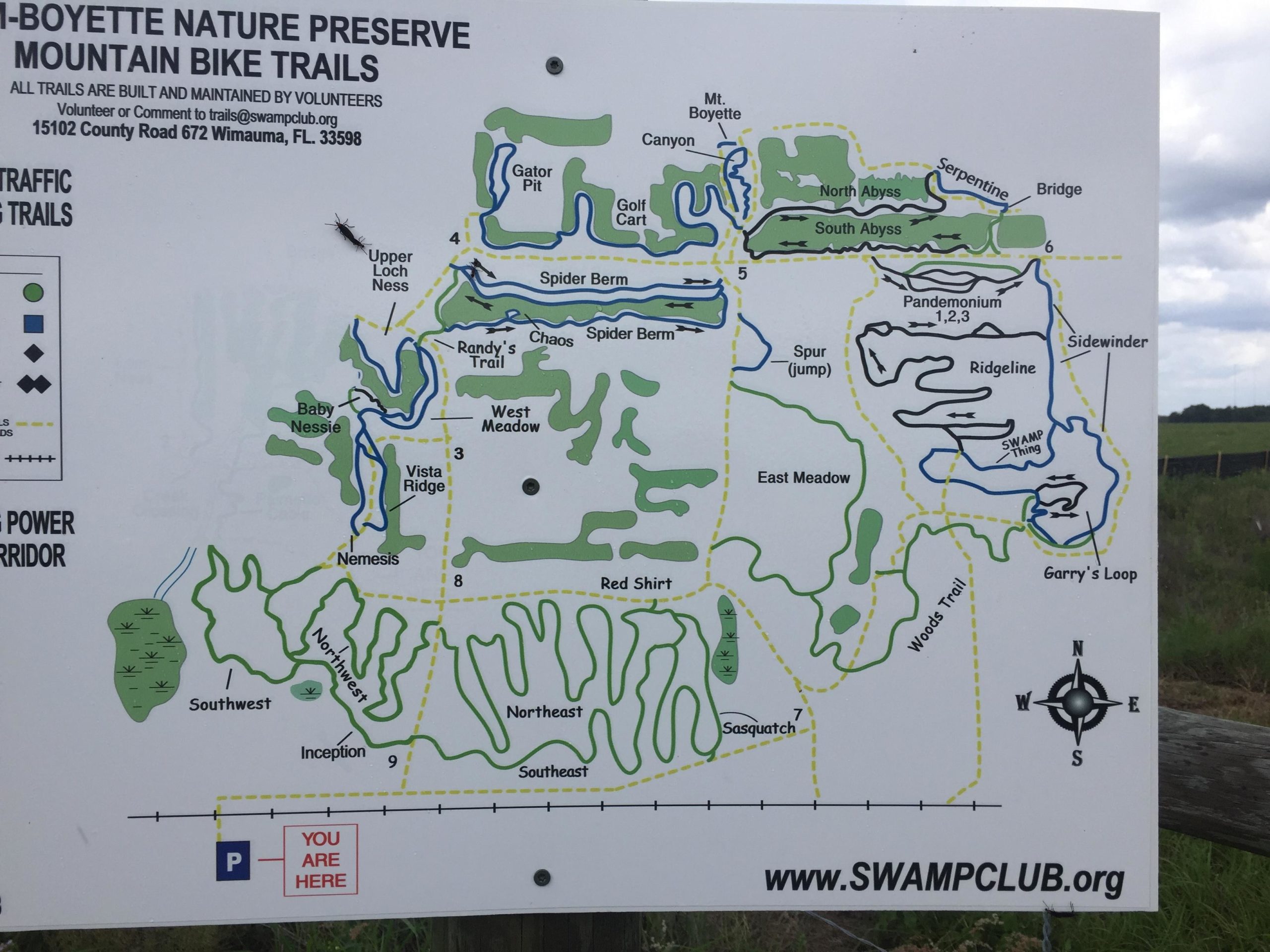 Map of the Boyette Nature Preserve mountain bike trails, featuring various trails such as "Upper Loch Ness," "Garry's Loop," and "Sasquatch." Includes marked paths, trail names, and a "You Are Here" indicator, along with a compass rose for orientation. The background consists of a grassy landscape under a cloudy sky. Balm Boyette Scrub Preserve mountain bike trail.