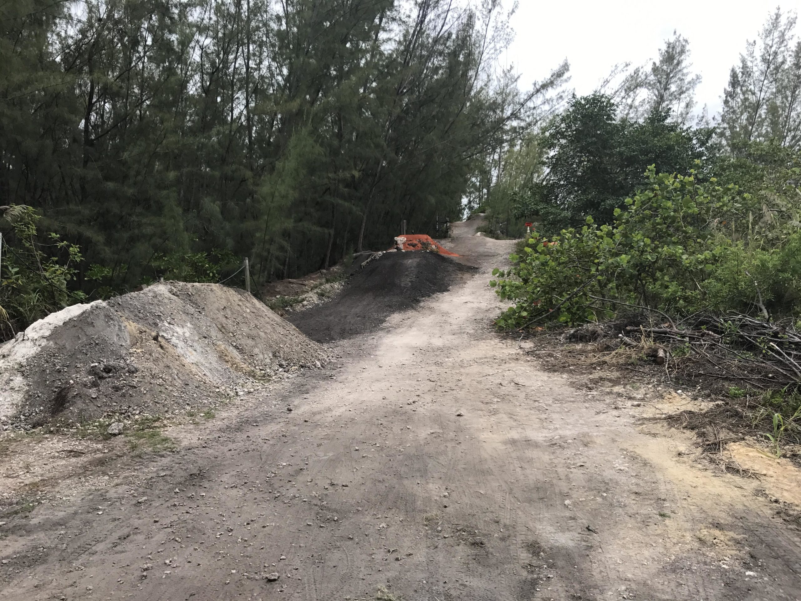 A dirt path leading into a wooded area, with piles of gravel and soil on either side. Sparse vegetation and trees are visible, and the sky appears overcast. The path shows signs of use, with tire tracks and loose gravel. Oleta River State Park mountain bike trail.