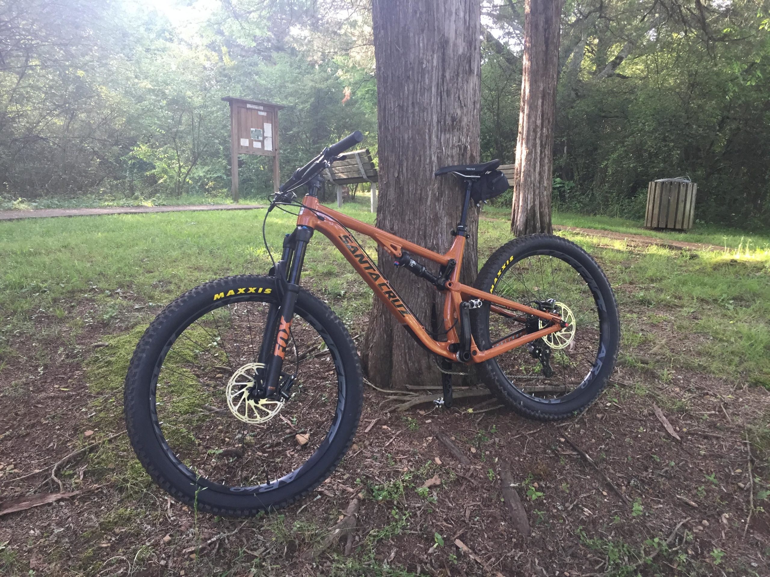 A mountain bike with an orange frame resting against a tree in a wooded area, with a trail visible in the background and a wooden information board nearby. The ground is covered with grass and leaves, and there is a trash bin in the distance. Shutes Branch mountain bike trail.
