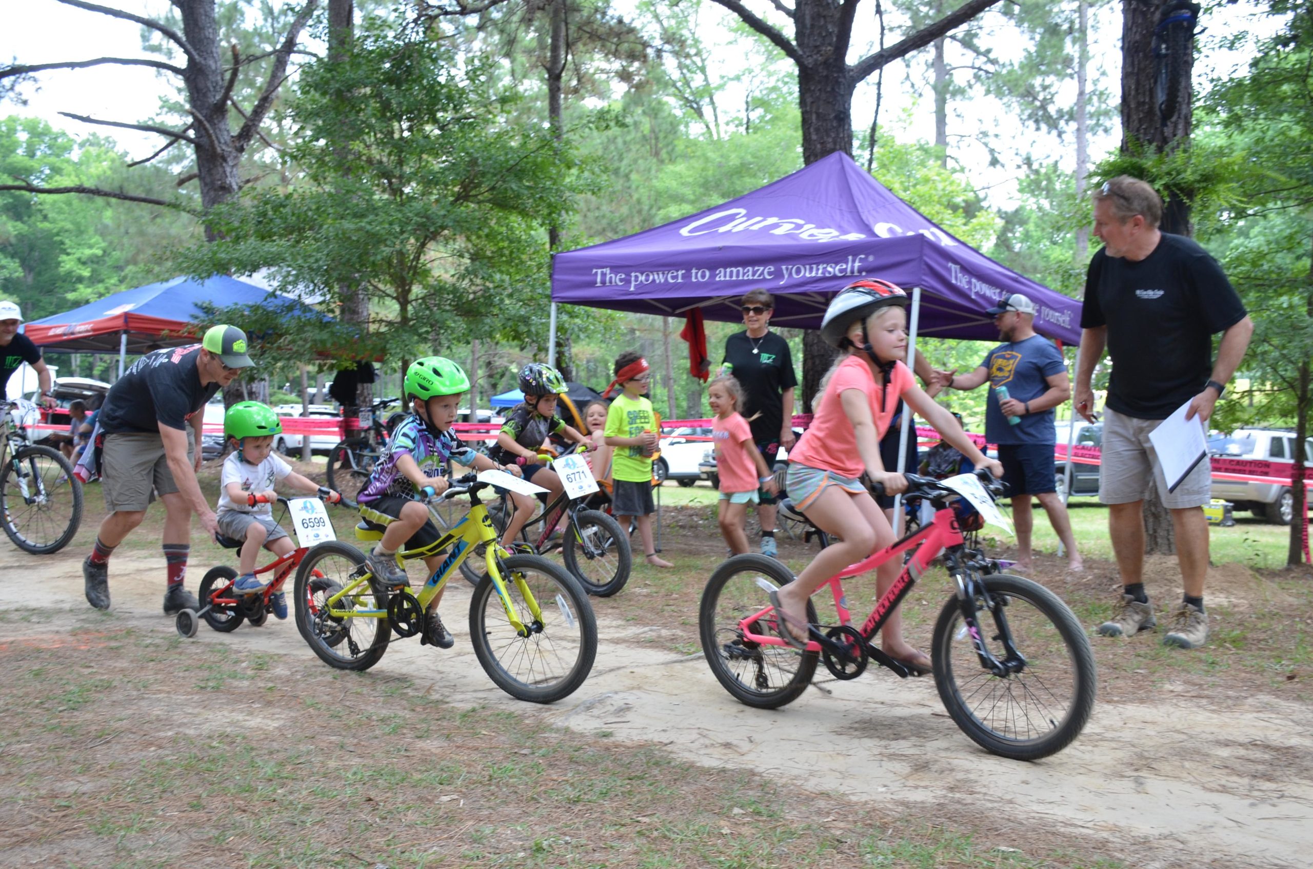 Children riding bikes in a wooded area during a race. Some kids wear helmets and race numbers, while an adult assists a younger child on a red bike. A purple tent is set up in the background, and several spectators watch from the side. Mt. Zion Bike Trails mountain bike trail.