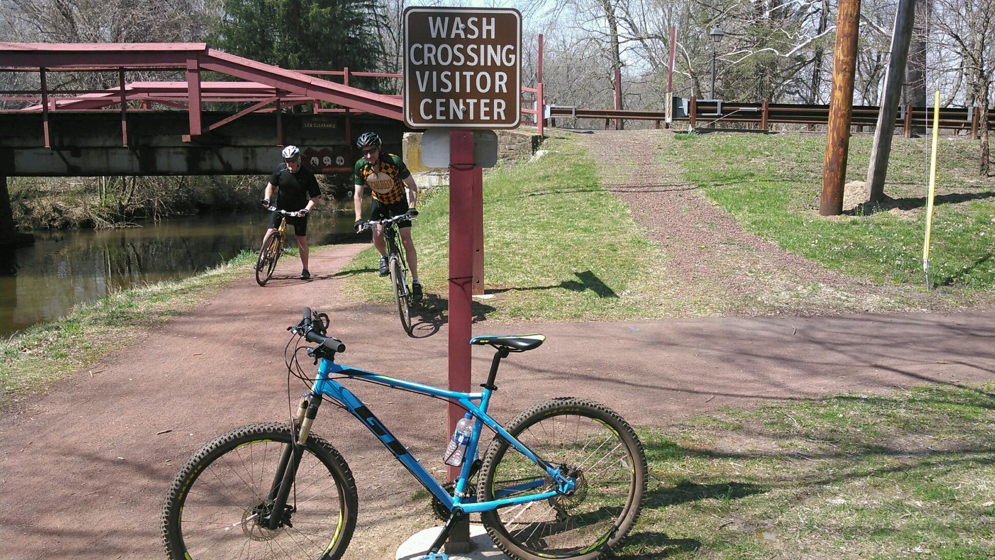 GT Aggressor Pro: A scenic outdoor path at Wash Crossing with a visitor center sign, featuring two cyclists near a stream. A blue mountain bike with a water bottle rests on the ground, while a red bridge crosses above. Trees and grassy areas are visible in the background, showcasing a pleasant spring day.