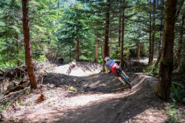Two mountain bikers navigate a dirt trail through a dense forest, surrounded by tall trees. The sunlight filters through the leaves, illuminating the path as one rider leans into a turn, while the other follows behind. Raging River State Forest mountain bike trail.