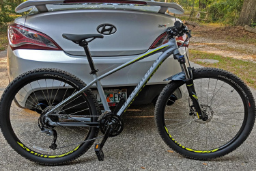 Specialized Pitch Comp: A gray mountain bike is parked next to a silver car, showcasing its sturdy frame and knobby tires. The bike's design features bright yellow accents, with the model name visible on the frame. The background includes trees and a gravel surface.