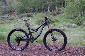 Pivot Mach 4: A mountain bike resting on a pine needle-covered ground, with a lush green forest in the background. The bike features a sleek design, thick tires, and a modern frame, suggesting it is built for off-road biking.