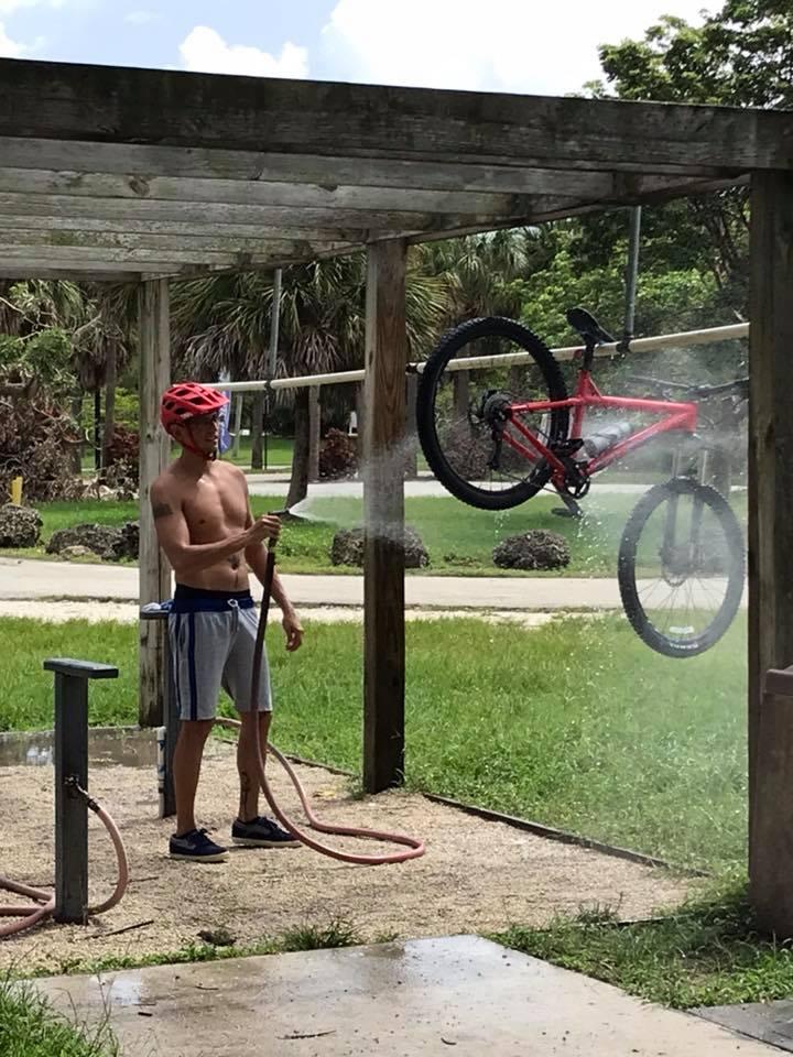 A person in a red helmet and shorts sprays water on a bicycle hanging from a wooden structure in a park. The scene features green grass and palm trees in the background, with a bright sky overhead. Quiet Waters Park mountain bike trail.
