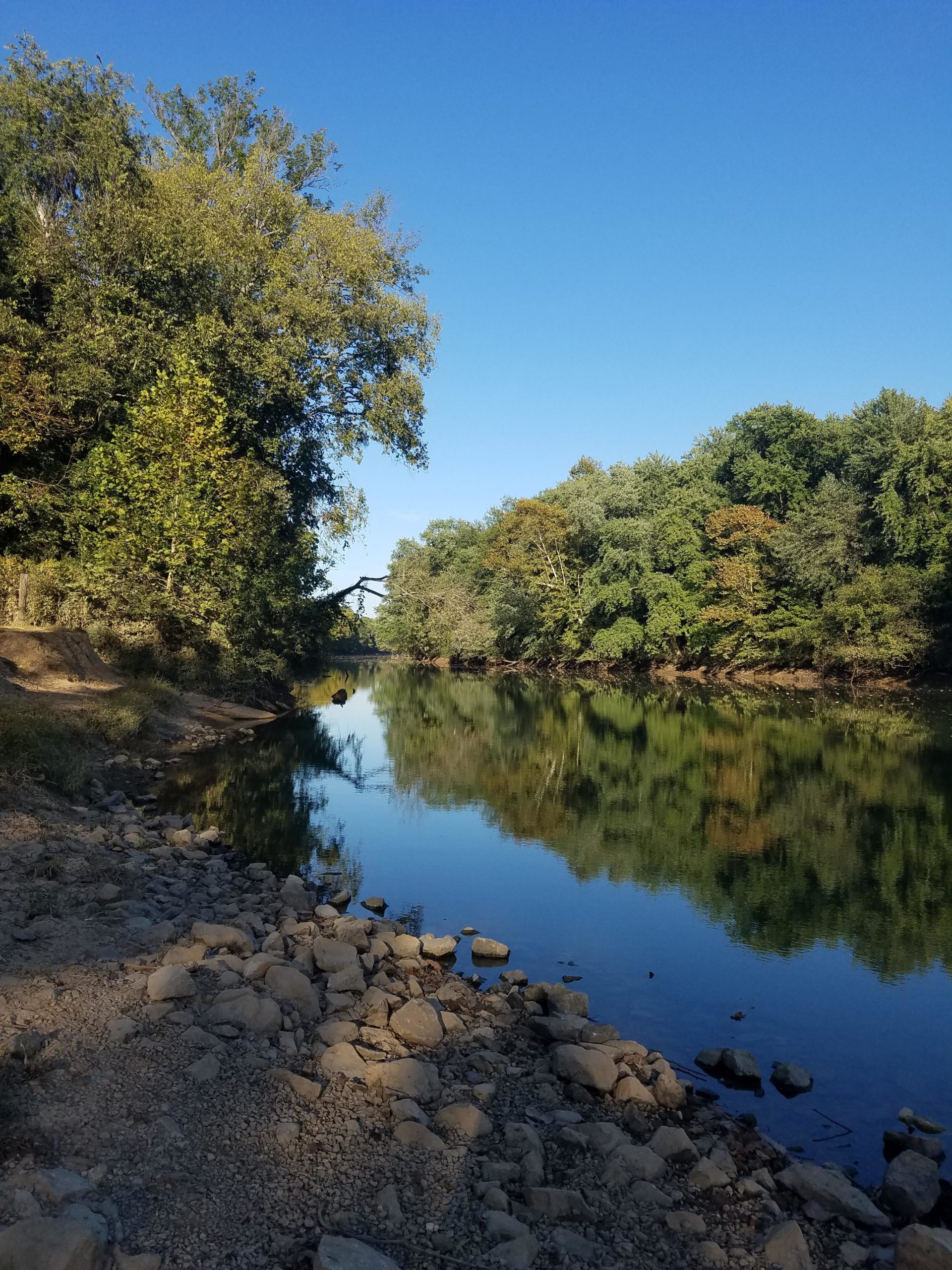 A tranquil river scene featuring clear blue skies and lush greenery along the banks. The water reflects the trees, creating a mirror-like effect. Pebbles and larger rocks line the river's edge, with a sandy path leading away in the background. Low Hollow mountain bike trail.