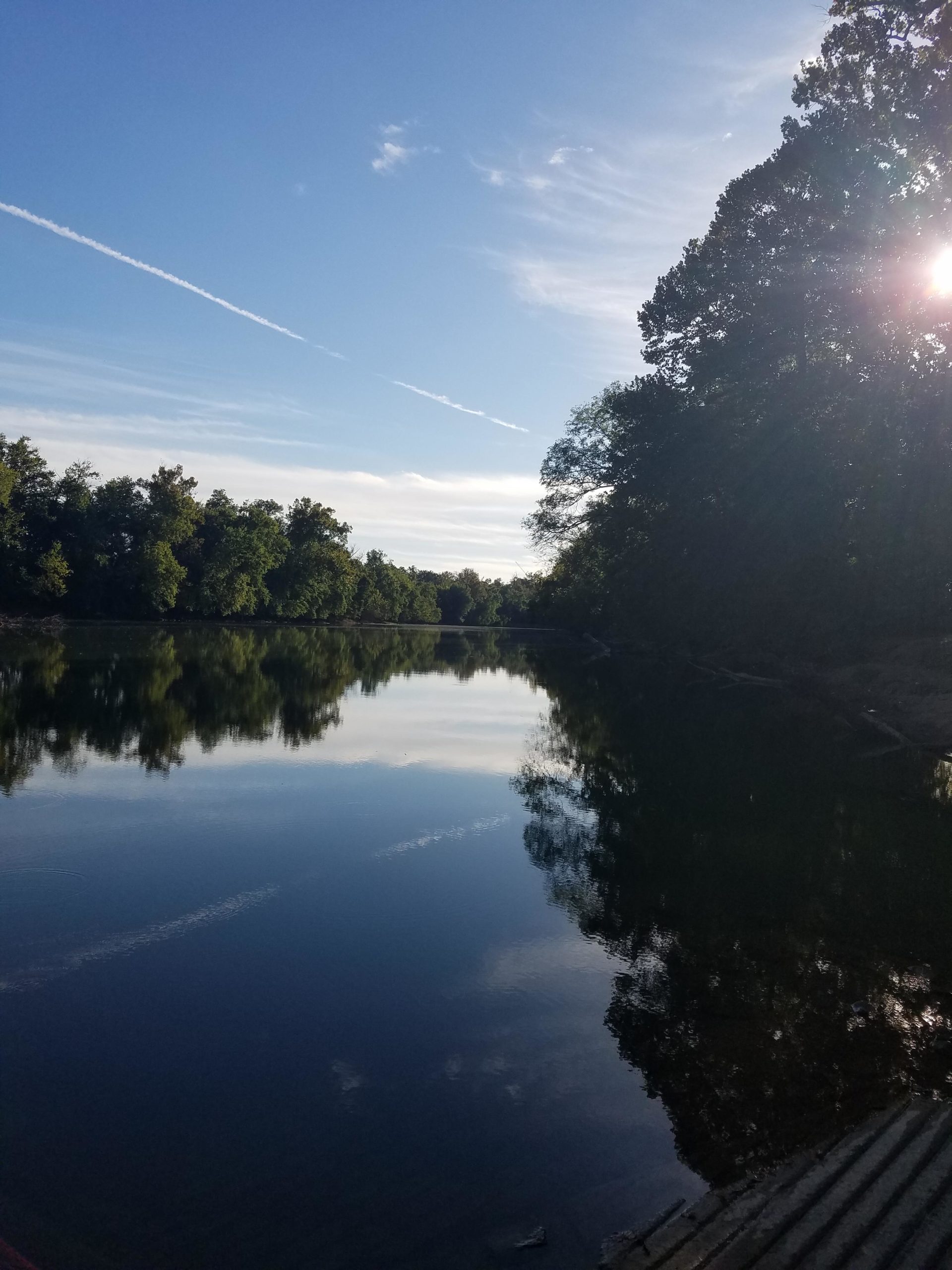 A serene view of a calm river reflecting the sky and surrounding trees, with soft clouds and a hint of sunlight peeking through the foliage. The scene captures a tranquil atmosphere, ideal for nature appreciation. Low Hollow mountain bike trail.