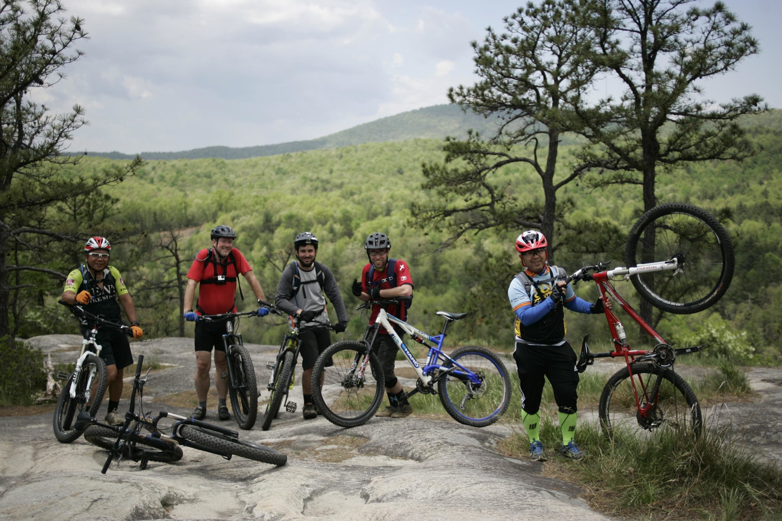 A group of five mountain bikers poses for a photo on a rocky outcrop surrounded by green trees and hills. One rider holds his bike above his head, while others stand next to their bicycles, smiling and enjoying the outdoor setting. The sky is partly cloudy, and the scene captures the spirit of adventure and camaraderie among the cyclists. Big Rock Trail #3 mountain bike trail.