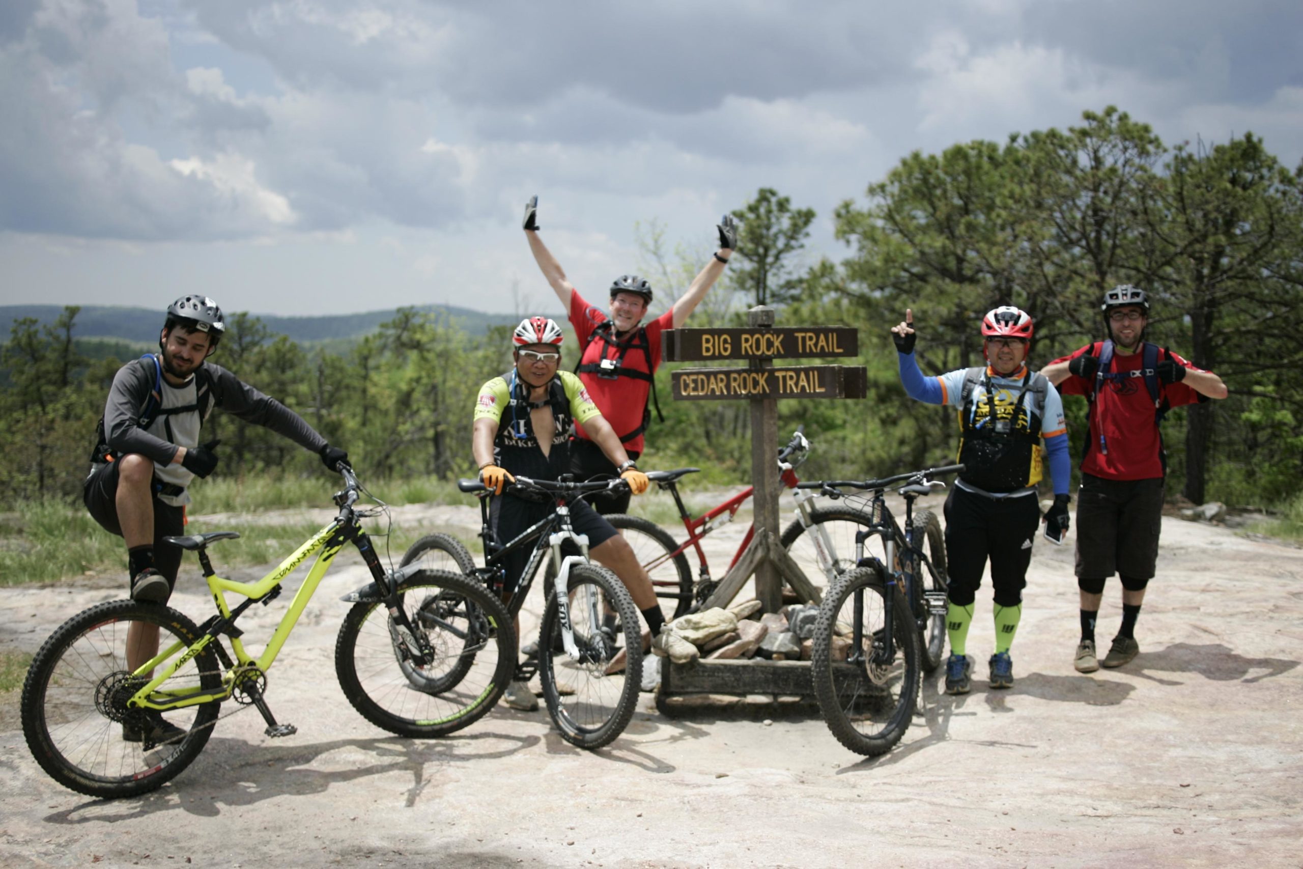 A group of five mountain bikers posing joyfully at a trail sign marked "Big Rock Trail" and "Cedar Rock Trail." They are wearing helmets and cycling gear, standing next to their bikes on a rocky trail surrounded by greenery and hills under a partly cloudy sky. Big Rock Trail #3 mountain bike trail.