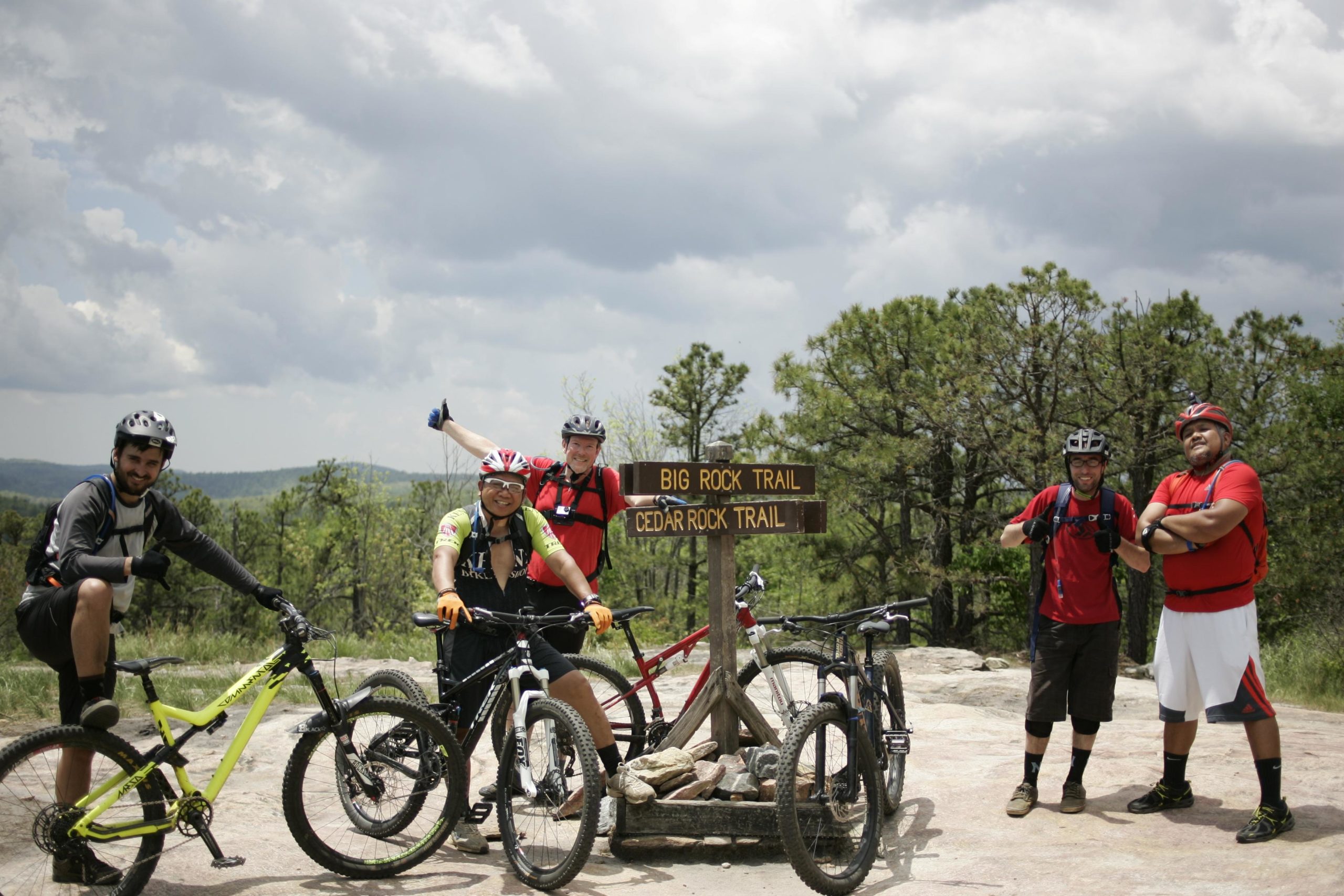 A group of five mountain bikers poses at a trail sign for Big Rock Trail and Cedar Rock Trail, surrounded by lush greenery and cloudy skies. They are wearing helmets and biking gear, with various mountain bikes parked nearby. The atmosphere is energetic and playful, showcasing the camaraderie of outdoor biking enthusiasts. Big Rock Trail #3 mountain bike trail.
