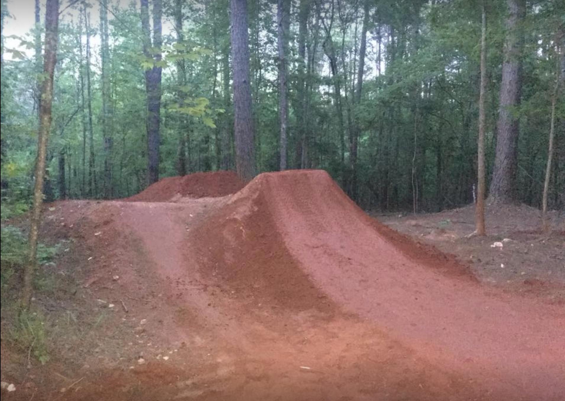A dirt ramp designed for biking, located in a wooded area. The ramp features two large mounds with smooth surfaces, surrounded by tall trees and lush greenery. The scene is set in a natural outdoor environment, suggesting a recreational space for cyclists. Blankets Creek mountain bike trail.